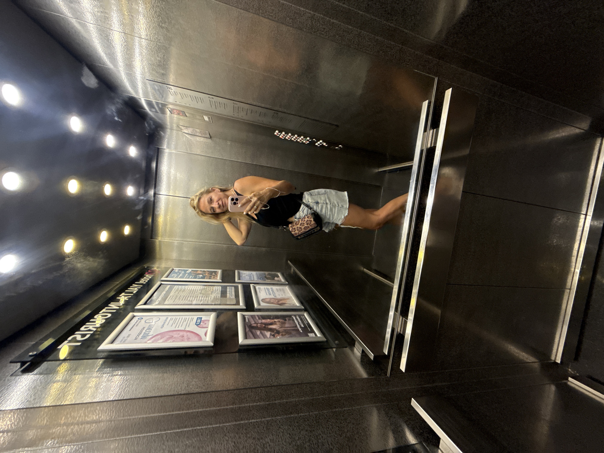 In an elevator with brushed metal walls, a woman with blonde hair and a black tank top and denim shorts takes a mirror selfie with her phone. She is wearing a leopard-print crossbody bag. The elevator's control panel is visible on the right wall, featuring a row of lit buttons. On the left wall, several framed pictures are displayed. The scene suggests the woman is in an indoor setting, possibly a hotel or residential building, in Yalta, Ukraine, given the sign with Cyrillic text on the left wall which reads "ГОСТИНИЦА 'ЯЛТА'".