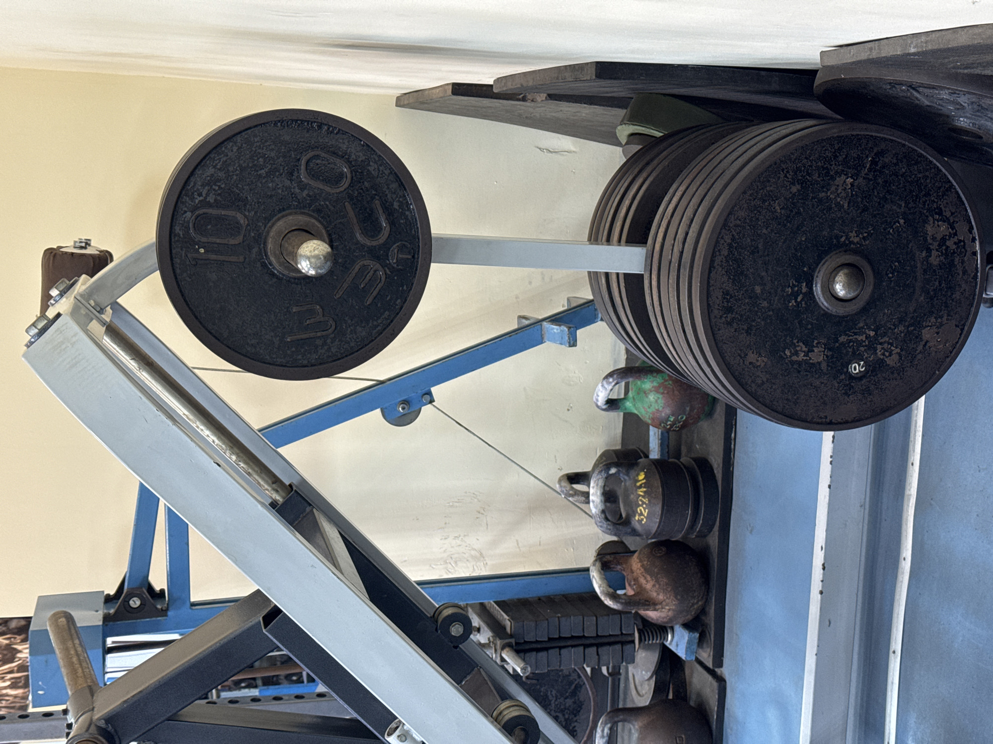 The image depicts a weightlifting setup, likely within a gym or home gym setting. A barbell is loaded with weight plates. On the left side of the barbell is a single 10-unit weight plate. On the right side are multiple weight plates stacked together, with a visible "20" marked on one. The setup includes a weight rack with blue and gray structural elements. Behind the weights and rack are several kettlebells of varying sizes and colors. The background is a light-colored wall. The city of Kladovitsy, Russia is not discernable from the image.