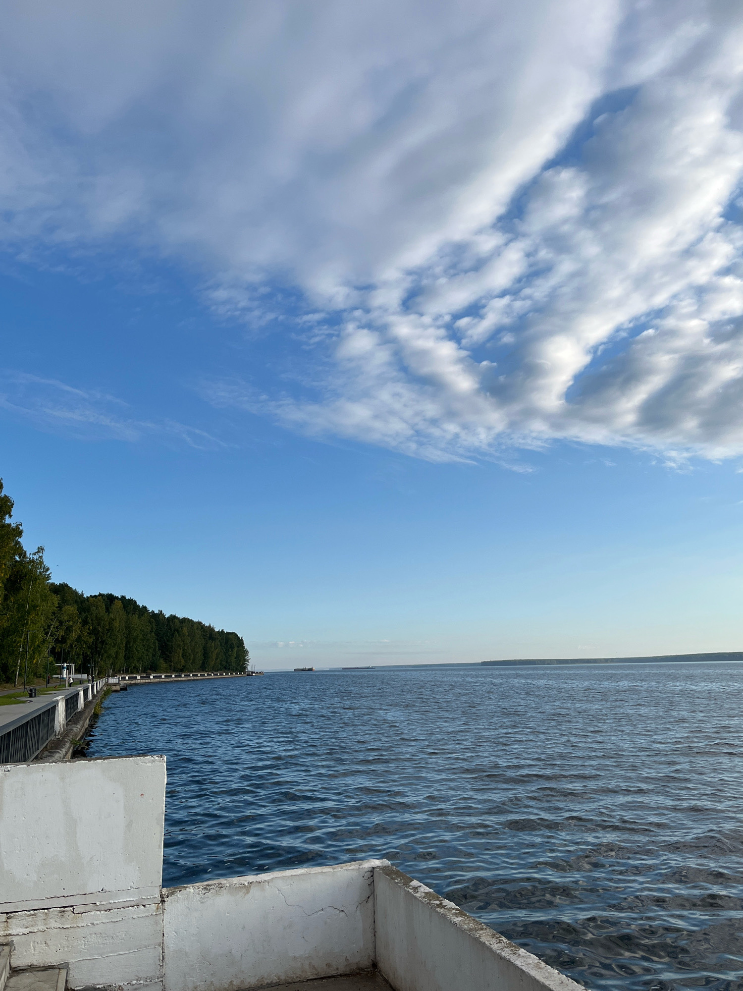 The image depicts a serene waterfront scene in the city of Chaikovsky, Russia. In the foreground, a weathered concrete barrier with white paint frames the view. Beyond this barrier, the calm, rippling blue water of a large body of water stretches out to the horizon. Along the left bank, a lush green treeline of deciduous trees lines a paved promenade, separated from the water by a dark metal railing. A few distant ships are visible on the water. The sky above is a vibrant blue, partially filled with dramatic, wispy white clouds.