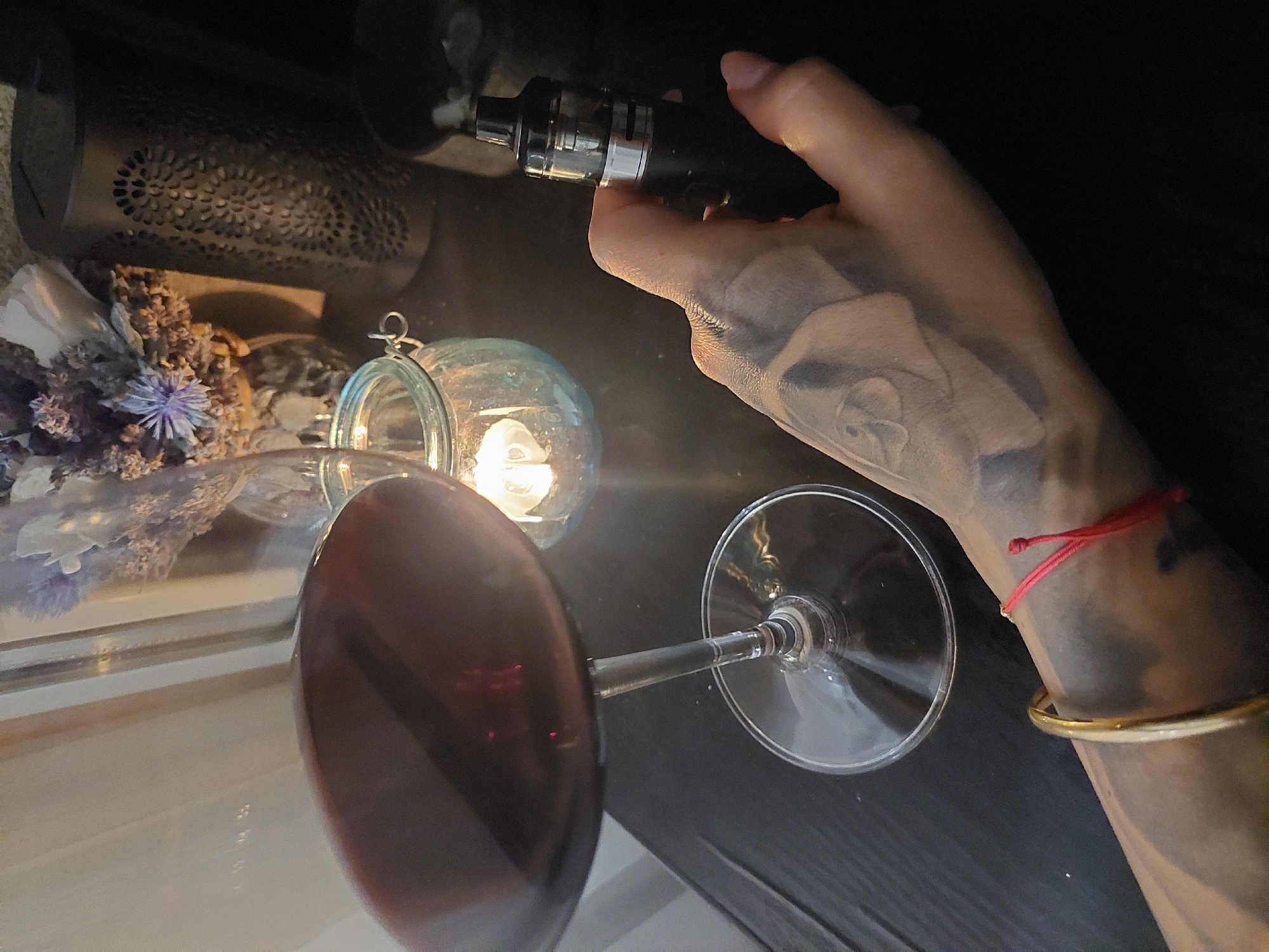 A close-up, high-angle shot depicts a hand holding a black vape pen over a wine glass filled with red wine. The hand has tattoos and is adorned with a red string bracelet and a gold bangle. The background includes a decorative metal lantern and a glass orb containing a light source, positioned near dried flowers. The location cannot be determined from the image alone, and I cannot confirm whether it is in Plovdiv, Bulgaria.