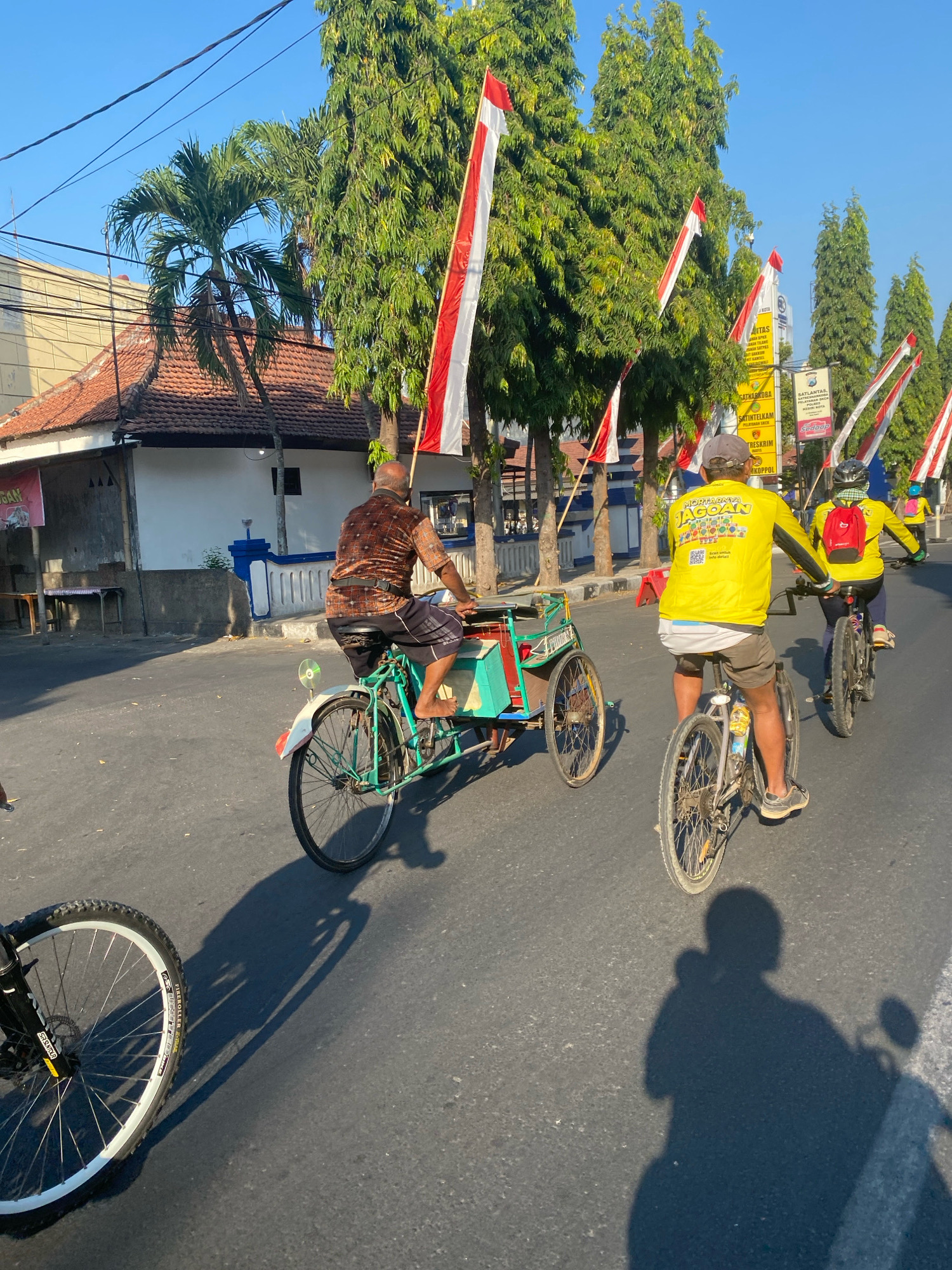The image captures a sunny day in Kediri City, Indonesia, with several individuals cycling along a paved street. In the foreground, a bicycle wheel and its shadow are prominently featured. Further down the street, a man in a brown and orange checkered shirt rides a green and teal pedicab, his bare feet visible. Behind him, two individuals in bright yellow long-sleeved shirts are on bicycles. The person in the middle wears a grey cap and the one behind them sports a black helmet and a red backpack. Several tall trees line the street, adorned with Indonesian flags. The background reveals buildings with red tiled roofs and a clear blue sky. The overall scene suggests a community event or casual cycling activity in an urban Indonesian setting.