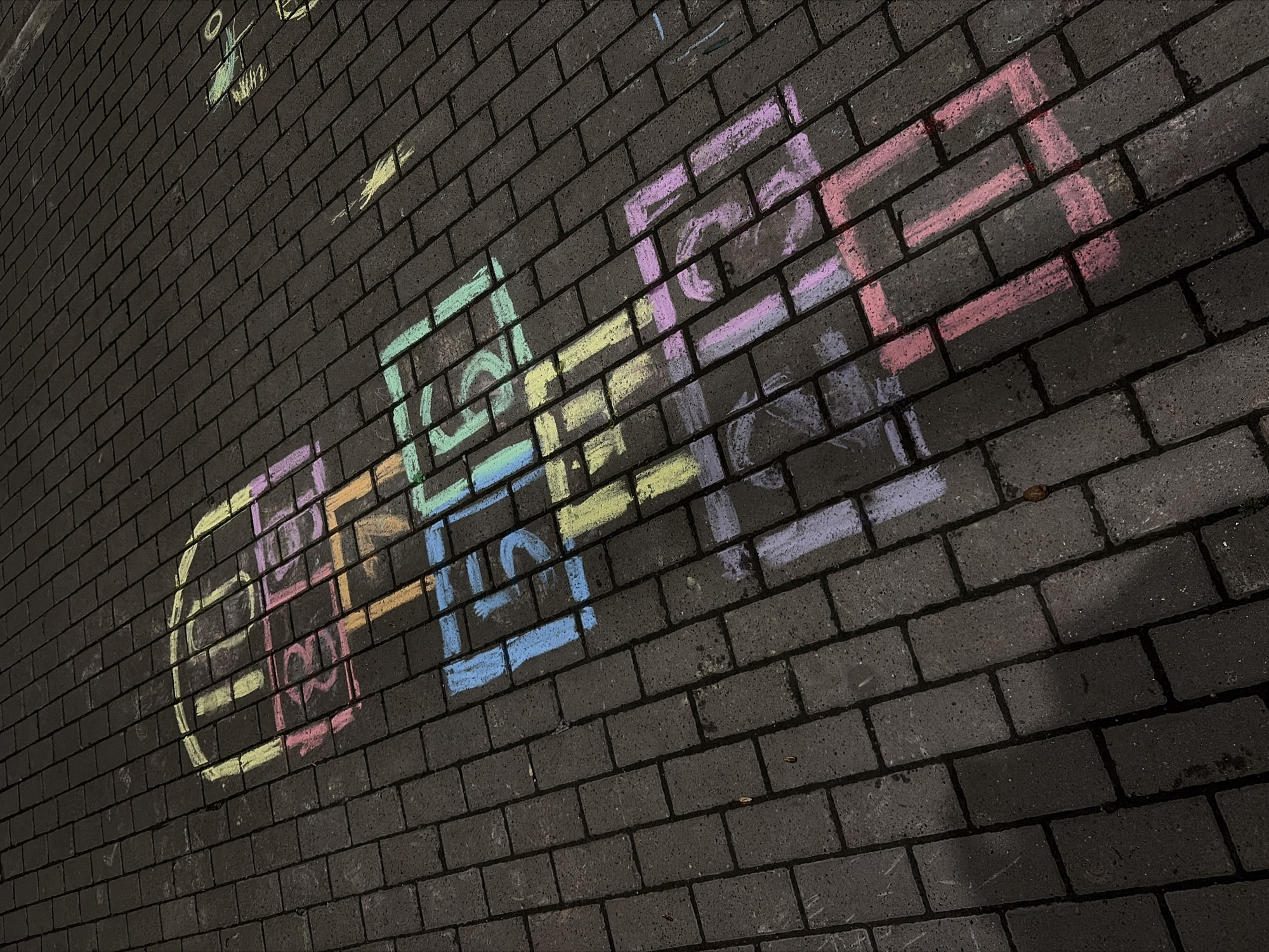 On a dark brick surface, a hopscotch court is drawn in colored chalk. The court, consisting of a circle, followed by single squares labeled "1," "2," "3," and then a split square with "4" and "5," concludes with a final square marked "6." The chalk colors are varied and include pink, purple, blue, yellow, and green. The brick surface appears to be a sidewalk or paved area. The location is Unknown, Unknown.