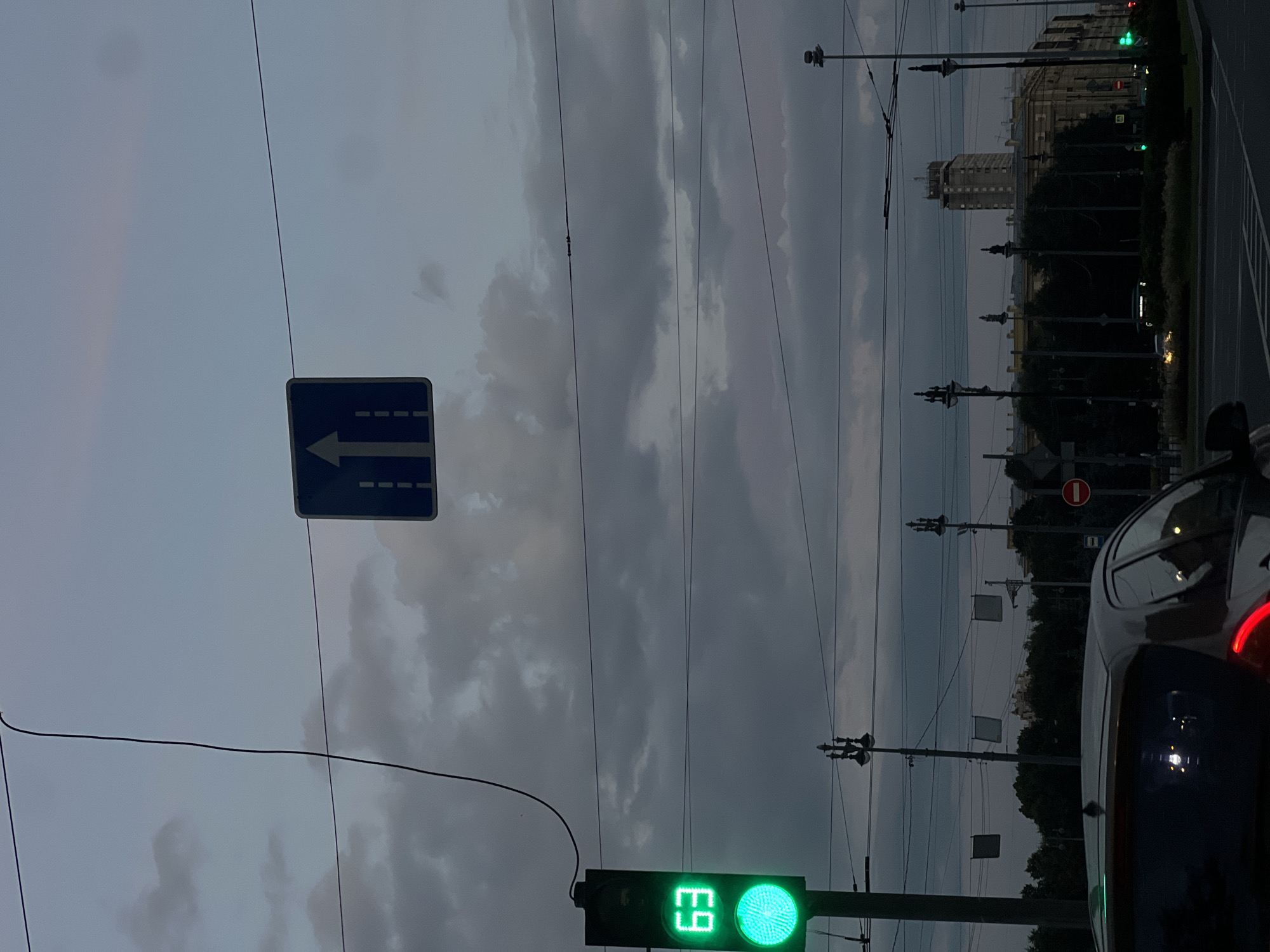 In Saint Petersburg, Russia, under a cloudy, twilight sky, a traffic light displays a green signal and the number "63". A blue road sign with a white arrow indicating a straight direction is suspended by wires overhead. The scene captures a street at dusk, with utility wires crisscrossing the sky, streetlights with ornate tops, and buildings visible in the background, including a tall, light-colored structure. A car is partially visible in the foreground, its rear window reflecting lights. The overall atmosphere suggests an urban environment preparing for nightfall.