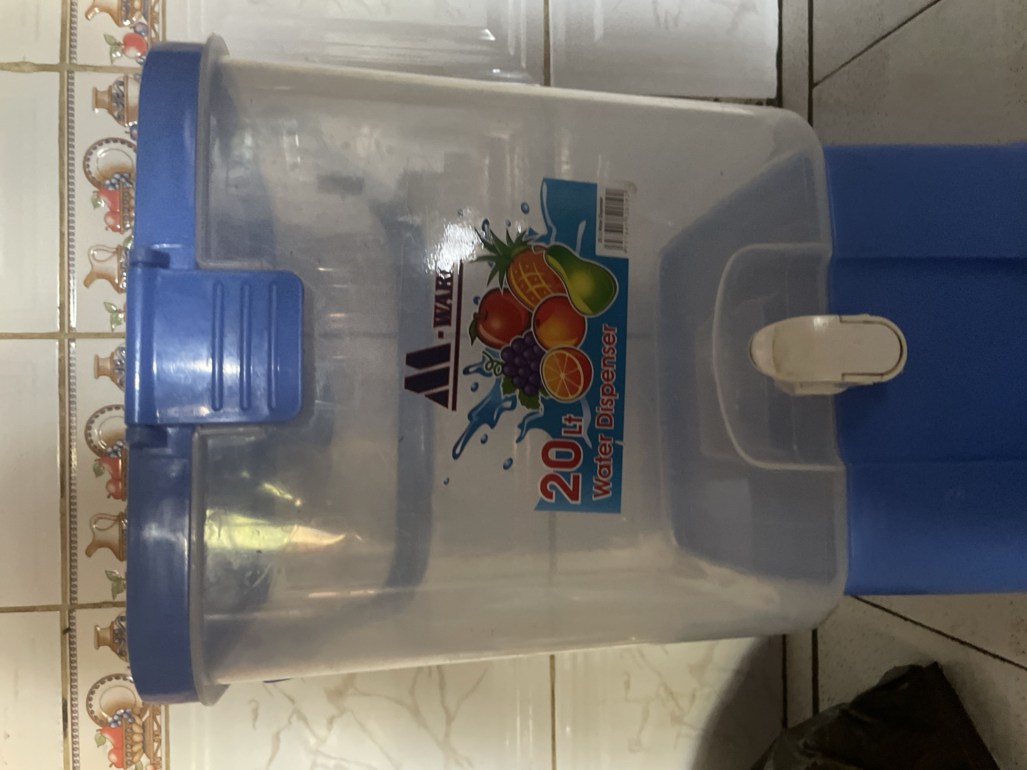 A 20-liter clear plastic water dispenser with a blue lid and base is shown in a kitchen. The dispenser has a decal on the front with fruit graphics and the text "20 Lt Water Dispenser". A white spigot is visible at the bottom. The dispenser is placed on a tiled countertop in what appears to be a residential setting. The background shows a tiled wall with a decorative border. The location is identified as Kakau, Nigeria. There are no people or actions depicted in the image.