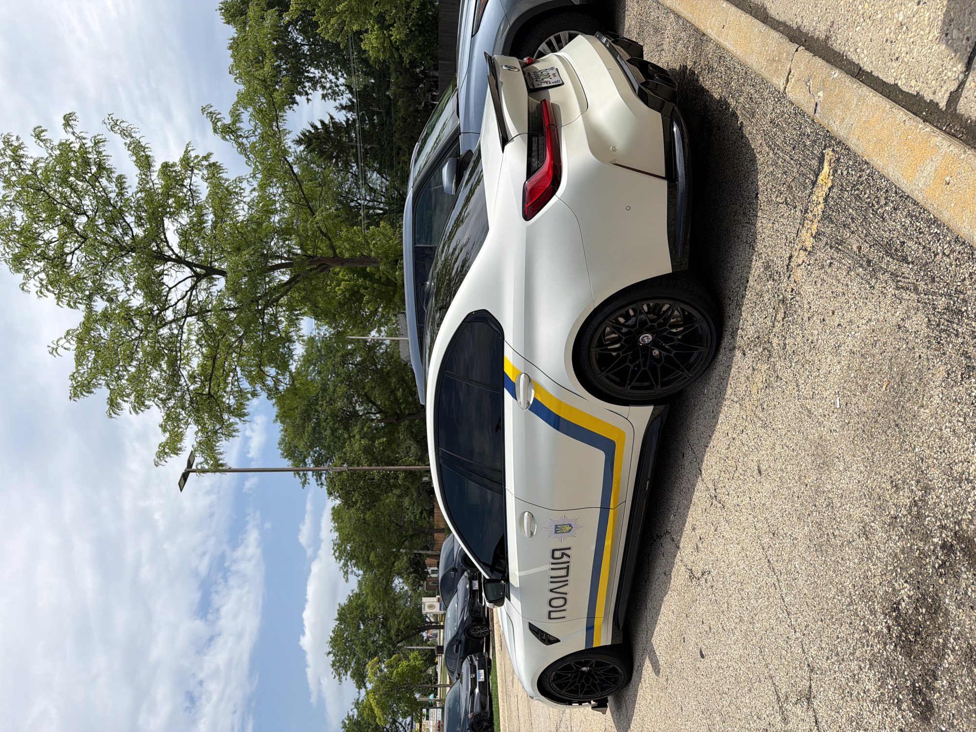 A white BMW M3 sedan, marked with "ПОЛІЦІЯ" in black lettering and a Ukrainian trident emblem, is parked in a lot in Buffalo Grove, Illinois. The car features blue and yellow stripes along its side, a black rear spoiler, and black wheels. Behind the BMW, a light blue SUV is parked. The scene is set outdoors on a paved lot with trees and a cloudy sky in the background.
