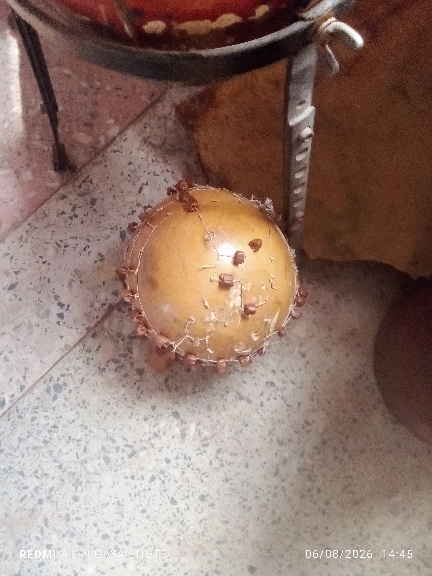 A close-up, high-angle shot depicts a spherical, light-brown object, possibly a gourd, adorned with cloves and wrapped in a delicate string net. The object rests on a speckled grey floor tile. In the background, a blurry metallic stand and a tan surface are visible. The bottom of the image displays text in white: "REDMI A1+| GIWA SHOTS" and "06/08/2026 14:45". The scene appears to be indoors.