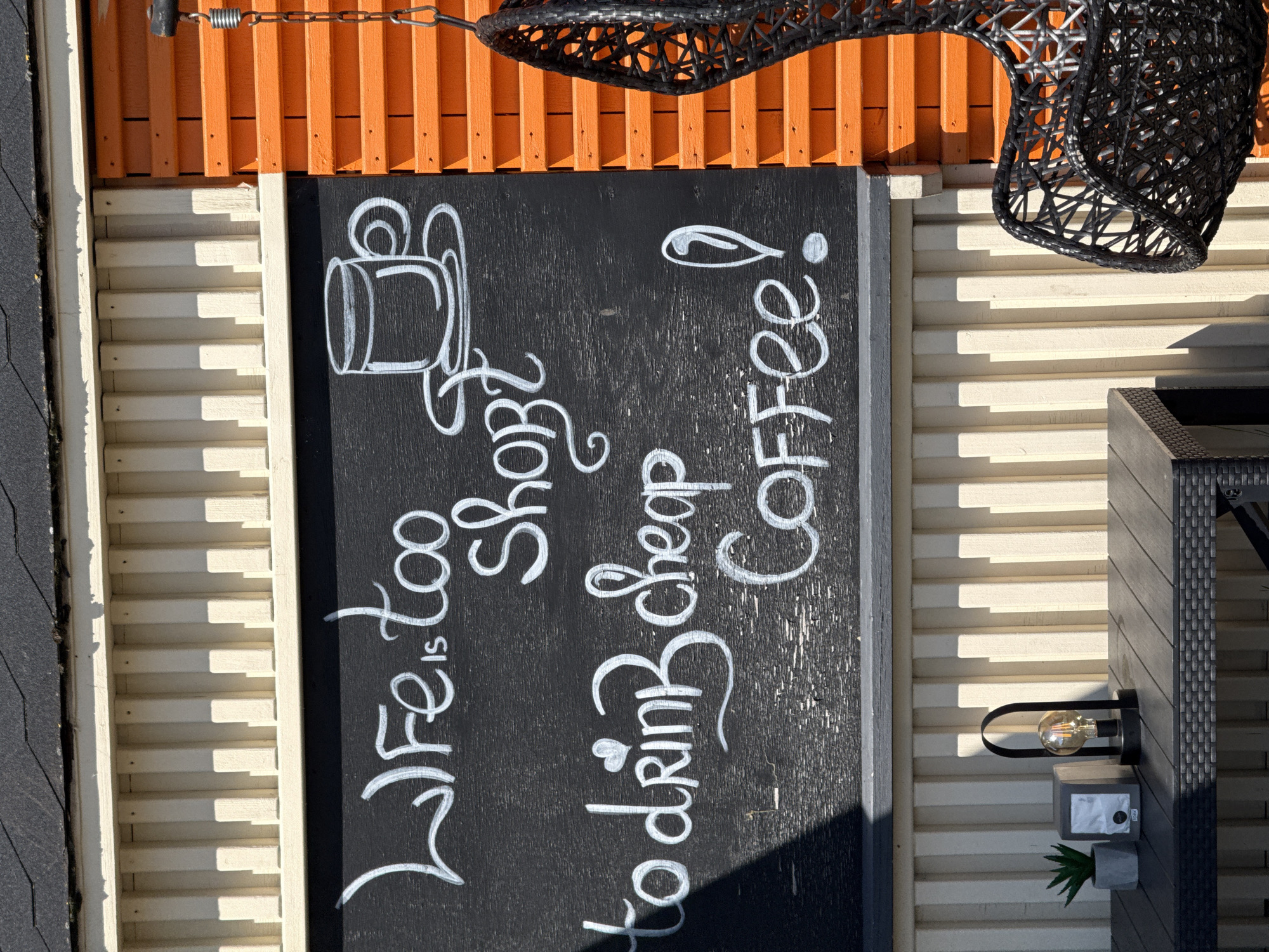 The image displays a close-up view of a rustic outdoor seating area, likely in Ventspils, Latvia, featuring a blackboard with white chalk lettering and drawings. The blackboard reads, "Life is too short to drink cheap Coffee!" and includes a drawing of a coffee cup and saucer. To the right of the blackboard, a portion of a bright orange, slatted wall is visible, with a black, woven hanging chair suspended by a chain and spring. In the lower left of the frame, a dark grey table is partially visible, with a small potted plant, a napkin dispenser, and a decorative lamp with a visible filament bulb resting on it. The background consists of light-colored, vertical wooden slats. The scene is illuminated by natural sunlight, casting shadows across the blackboard and table.