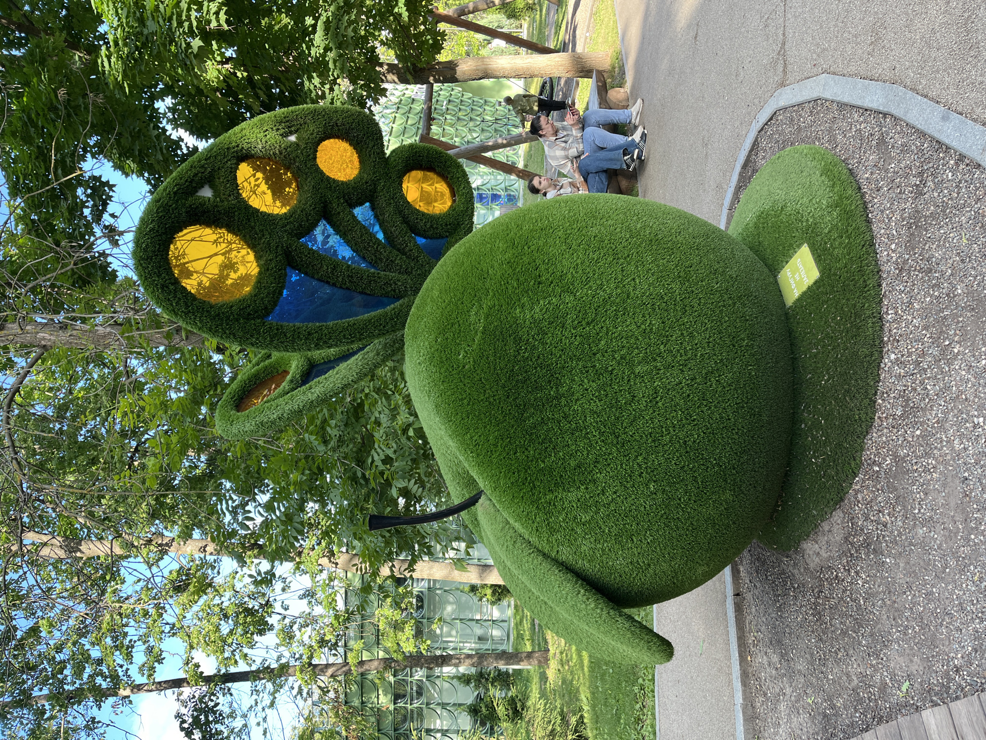 An oversized, artificial grass sculpture resembling an apple with butterfly wings is the central focus of this image, situated in a park in Nizhny Novgorod, Russia. The apple is a vibrant green and has a black stem. The butterfly wings are adorned with translucent yellow and blue panes. Two young women are seated on a wooden bench in the background, both looking at their phones. One woman wears a plaid shirt and jeans, while the other has a lighter colored plaid shirt and jeans. Behind them, a person in a green jacket stands near a wooden swing set. The environment appears to be a well-maintained park with trees, grass, and paved pathways. A small yellow sign with Cyrillic text is visible at the base of the sculpture.