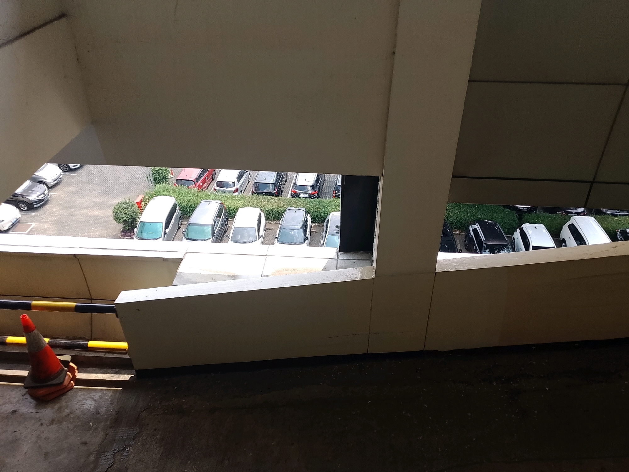 The image captures a view from an elevated concrete structure, likely a parking garage or overpass, looking down onto a parking lot filled with various vehicles. In the foreground, a red and orange traffic cone rests on a worn concrete surface, with a yellow and black striped barrier pole extending horizontally to the right.
Below the elevated structure, a row of cars is parked on a cobblestone-like surface. These include several white hatchbacks, a black SUV, and a red sedan. To the right of these vehicles, a row of parked cars is partially visible, including a white SUV, a black sedan, and several other vehicles obscured by the angle. A line of green bushes runs along the edge of the parking lot, separating it from what appears to be another section of pavement.
The scene is set in South Jakarta, Indonesia. The overcast sky and the parked vehicles suggest an urban environment, possibly an office building or commercial complex. There are no people visible in the image. The overall impression is one of a typical, functional urban parking area.