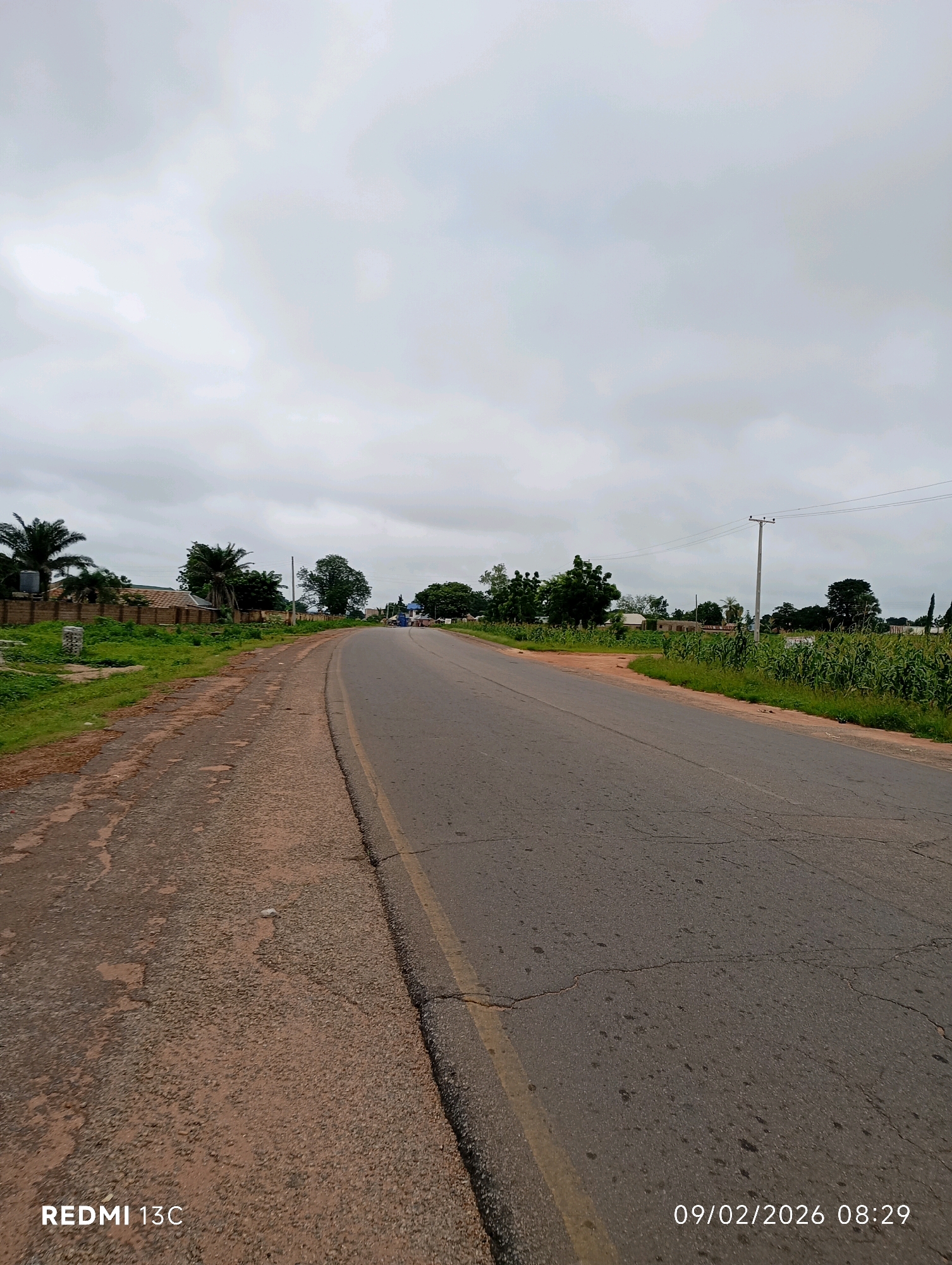 The image depicts an overcast day in Shendam, Nigeria, with a paved road curving through a semi-urban landscape. The road surface is grey asphalt, showing signs of wear and cracking, with a faded yellow line marking the center. To the left of the road, a rougher dirt path runs parallel, bordered by sparse, dry vegetation. Further left, a low brick wall separates the path from a green, grassy area with scattered small trees and some palm trees. Beyond the wall, residential buildings with tiled roofs are visible. To the right of the road, a strip of lush green vegetation, resembling tall crops like corn, stretches into the distance. A single utility pole with wires is present on the right side of the road. The sky is uniformly gray with heavy cloud cover, suggesting a lack of direct sunlight. The overall impression is a quiet, possibly rural or suburban, area on a cloudy day, with evidence of ongoing construction or development visible in the background. A timestamp in the lower right corner indicates the date and time: 09/02/2026 08:29. The text "REDMI 13C" is present in the lower left corner, likely indicating the device used to capture the image. There are no people or animals visible in the frame.