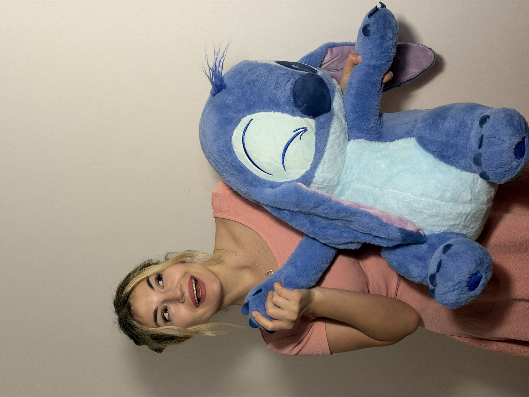 An indoor, eye-level shot captures a young woman playfully holding a large blue plush toy. The woman, with blonde highlights in her hair, is seen from the chest up, smiling broadly with her mouth open, revealing teeth. She is wearing a light pink, textured short-sleeved top and a delicate silver necklace with a circular pendant. Her head is tilted, and her gaze is directed upwards and to the left, conveying a sense of joy or amusement.
She is holding a large, soft blue plush toy, clearly identifiable as Stitch from Disney's Lilo & Stitch. The toy features lighter blue fur on its belly and inner ears. Its face shows embroidered sleepy eyes. The woman's hands are engaged with the toy; her left hand holds one of Stitch's feet, while her right hand is near its midsection. The overall impression is one of affectionate interaction with the toy.
The background is a plain, light beige or off-white wall, offering no specific location details. The lighting is soft and even, typical of indoor conditions, with no indication of time of day or weather. No visible text is present in the image. The scene suggests a lighthearted moment, taking place indoors in Budva, Montenegro.