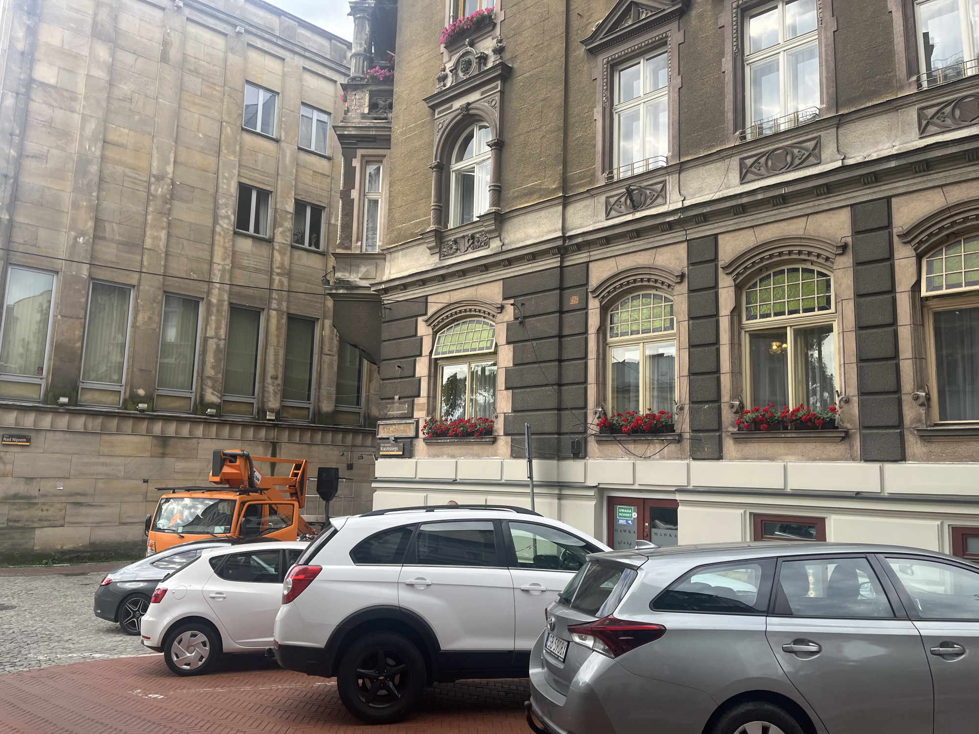 The image shows a street scene with buildings and cars in the city of Unknown, Unknown. On the left, a beige stone building with several windows of varying sizes is visible. To the right of it, a darker colored building with ornate architectural details and arched windows filled with flower boxes is prominent. In the foreground, several cars are parked. An orange cherry picker truck is parked behind a grey sedan and a white hatchback. The white hatchback is parked in front of a silver station wagon. The street is paved with cobblestones. The sky is overcast.
