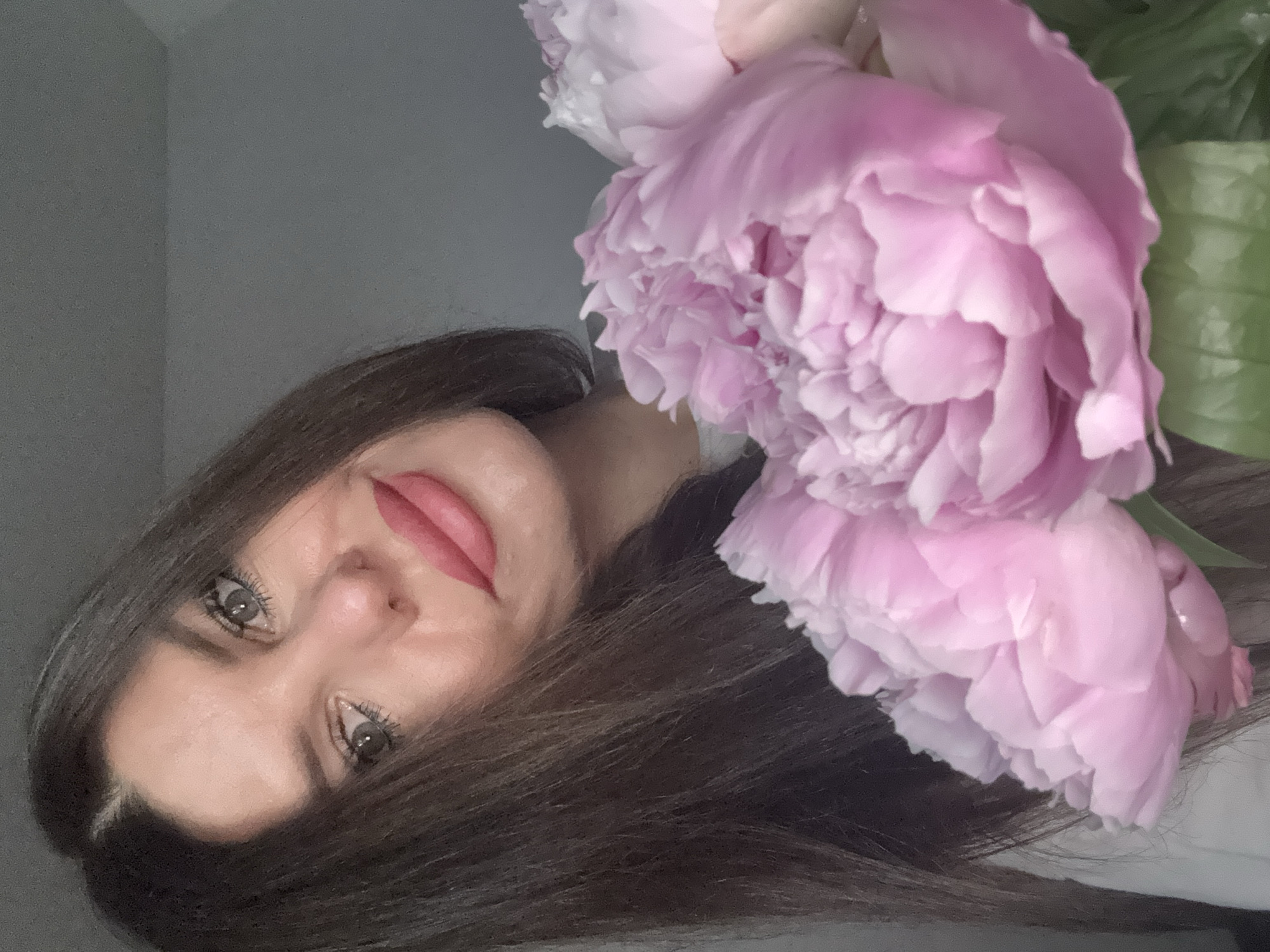 The image shows a close-up portrait of a woman holding a bouquet of pink peonies. She has dark brown hair and is wearing pink lipstick. The background is a plain grey wall. There is no indication of the city of Chaikovsky, Russia in the image.