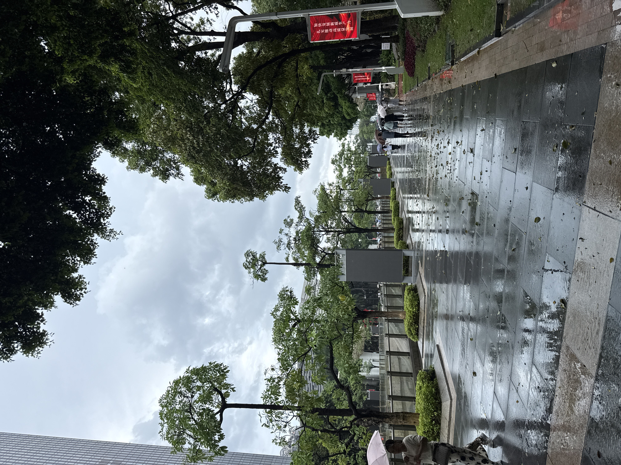 The image depicts a wet, tiled pedestrian walkway in Tianhe District, China, on an overcast day, likely during or after rainfall. Several individuals are walking along the path, many holding umbrellas. Trees with green foliage line the walkway on both sides, with one side featuring a modern building in the background. The wet surface of the walkway reflects the surrounding environment and fallen leaves are scattered across it. Red banners with Chinese characters are displayed on poles, suggesting a public event or advertisement. A single person in the foreground is walking with a pink umbrella. The scene conveys a sense of urban life experiencing inclement weather.