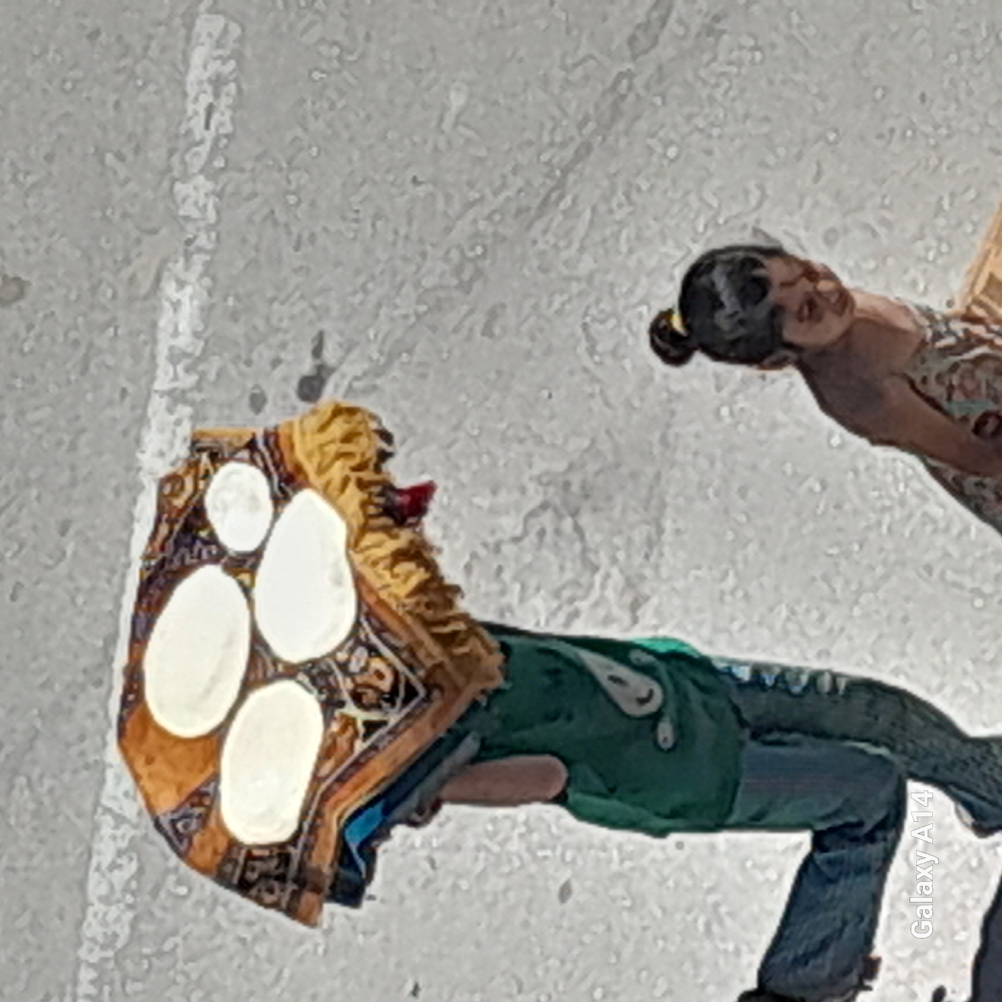 In Khan Yunis, Palestinian Territories, a young boy is carrying a stack of round, flatbreads on his head, covered by a patterned cloth. He is wearing a green shirt and dark pants. To his right, a younger girl, with her hair in a bun, is looking down as she walks. The ground appears to be a paved surface with some white markings. The image is taken from an elevated perspective.