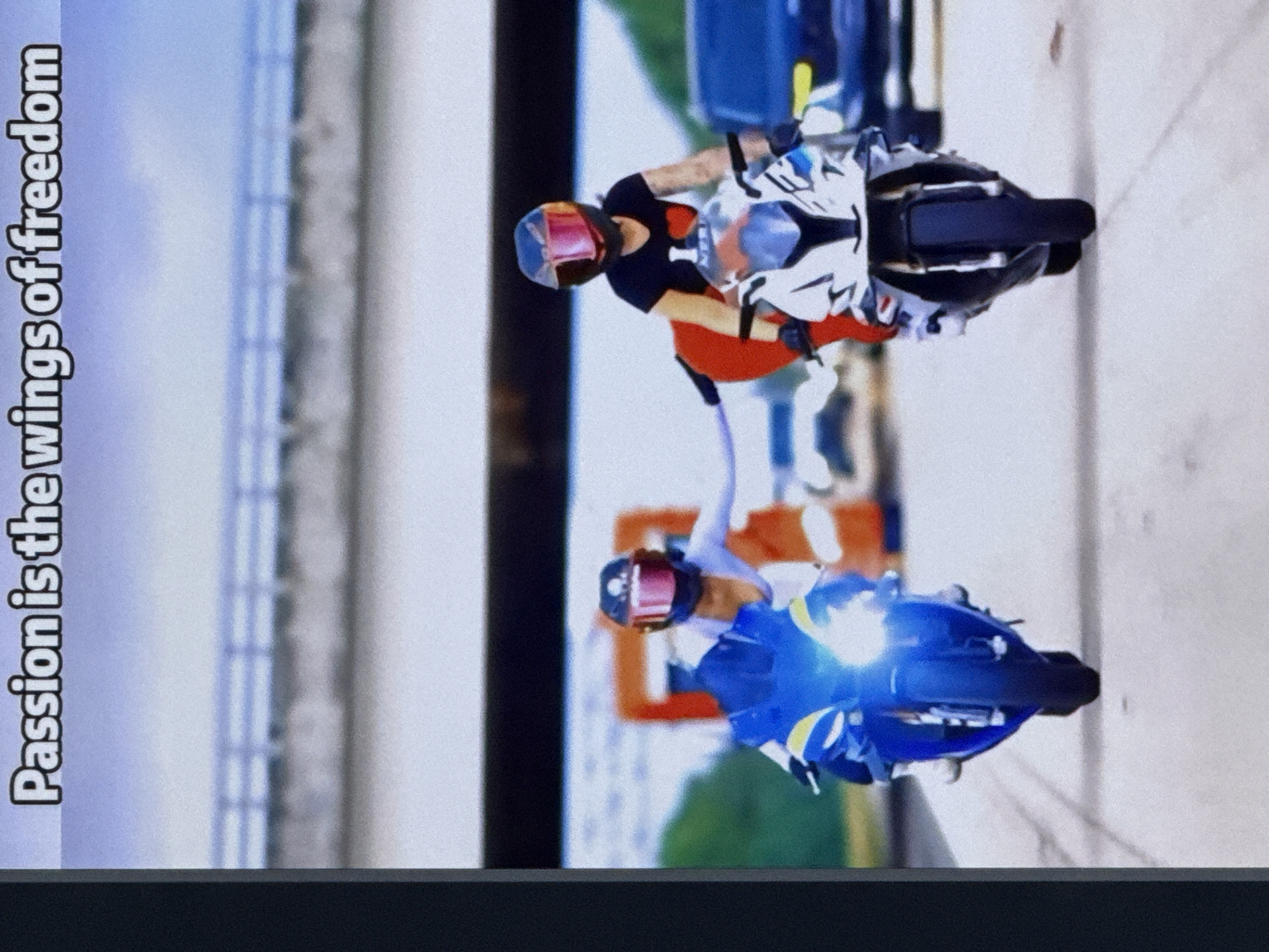 The image depicts two individuals riding sport motorcycles on a paved surface, likely a road or highway. Both riders are wearing helmets with red-tinted visors, and one rider on the right is wearing a black t-shirt with a red heart graphic. The motorcycle on the left is blue with yellow accents, and its headlights are illuminated. The motorcycle on the right is white with blue and silver accents. In the background, there are blurred orange traffic cones, a dark-colored vehicle, and a bridge or overpass structure. The sky is overcast, suggesting a daytime scene. The text "Passion is the wings of freedom" is overlaid at the top of the image. There is no visual information within the image that indicates the location as Cahul, Moldova.
