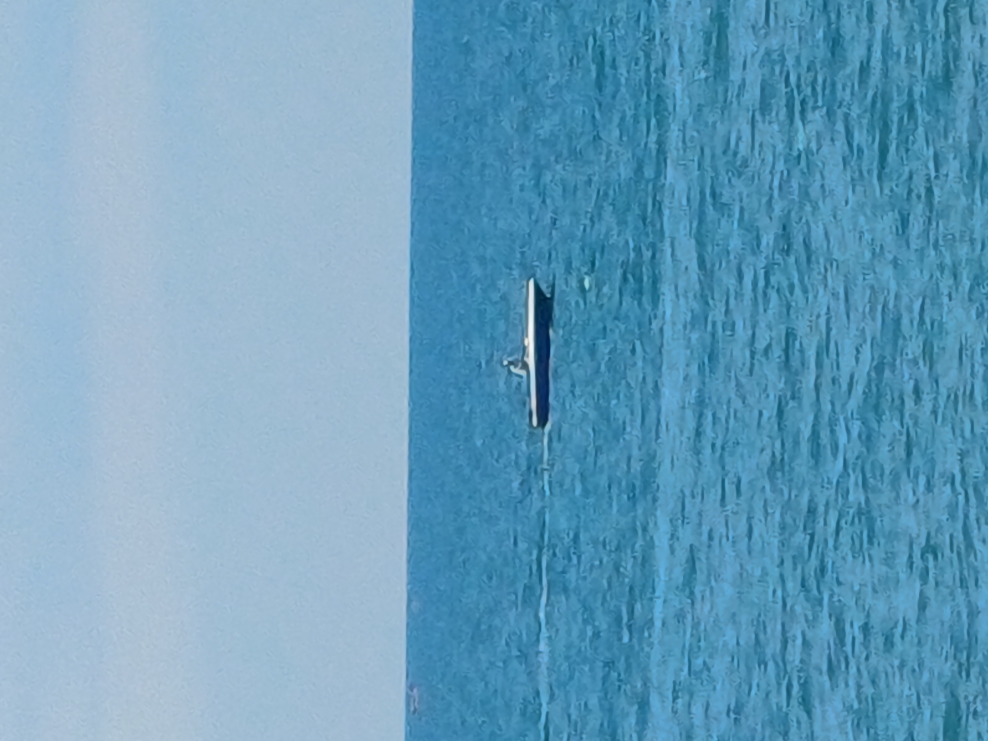 A small white and blue motorboat with one person on board is moving across a calm, blue sea under a hazy, light blue sky. The boat is leaving a slight wake behind it. In the far distance, another smaller boat is visible on the left. The scene appears to be a clear day at sea.