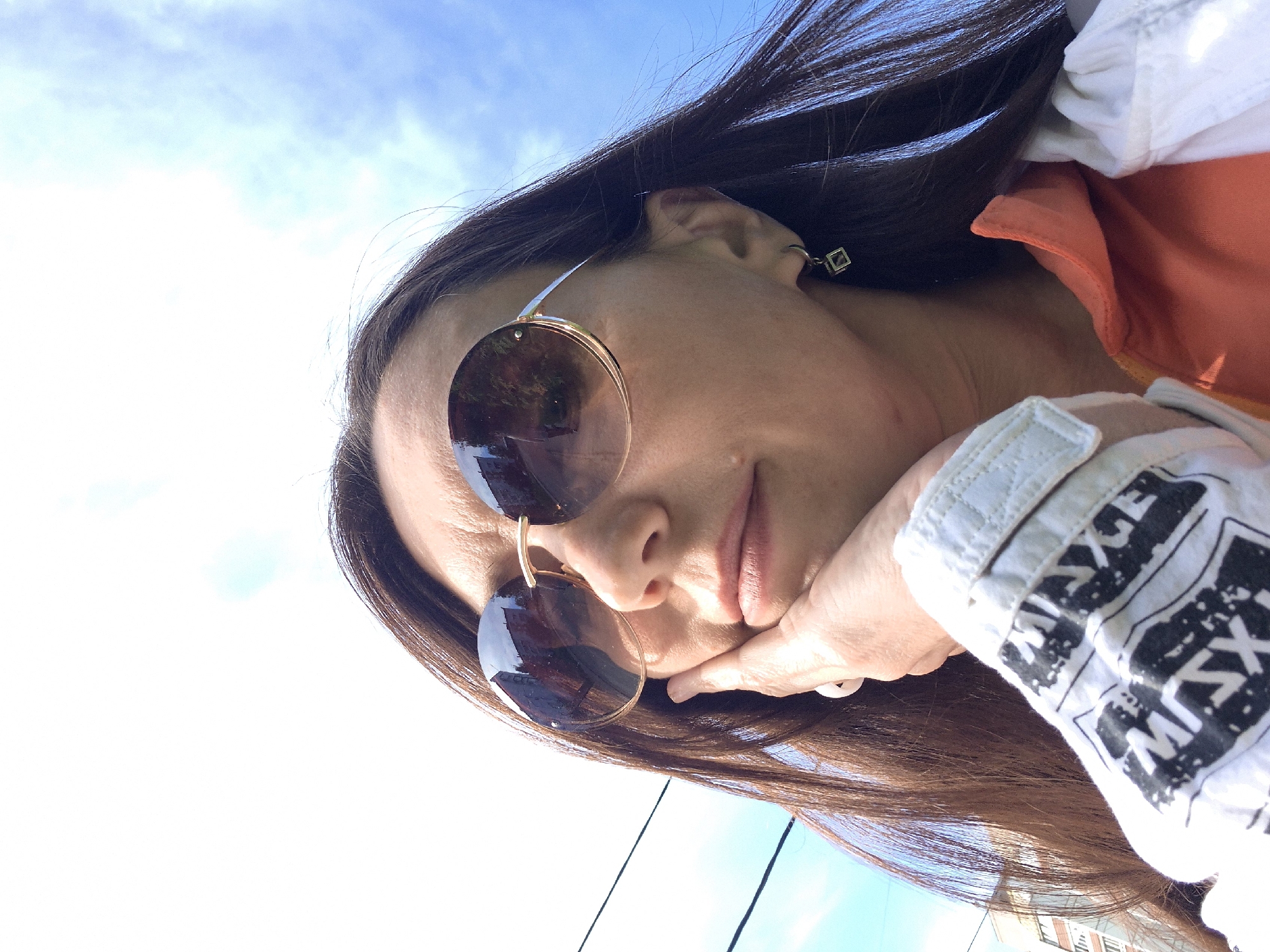 An angled, close-up outdoor shot features a woman with long, dark brown hair against a bright blue sky with scattered white clouds. She is looking slightly downwards and to her right, her chin resting on her hand. Her face is partially obscured by large, round, gold-rimmed sunglasses with reflective lenses, which show indistinct reflections of what appear to be buildings or trees.
She is wearing a white long-sleeved garment, possibly a jacket or shirt, that has bold black text patterns on the sleeve; partially visible words include "MAXGE" or "MXG" and "MIX" or "MXS". An orange piece of fabric, possibly part of another layer of clothing or lining, is visible beneath the white garment. A small, square, gold-colored earring can be seen in her right ear.
The background is dominated by a clear, bright blue sky, indicating a sunny daytime setting. A few thin black lines, likely power lines or cables, are visible against the sky in the lower left quadrant. The overall scene suggests a casual outdoor moment in an urban or suburban environment, potentially in Chaikovsky, Russia.