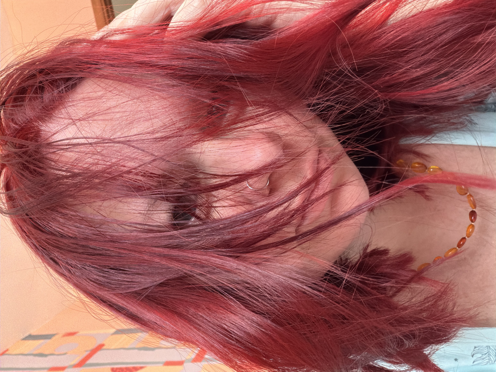 The scene is an interior setting within Chaikovsky, Russia, depicting a close-up portrait of a single individual. The subject is a female with long, vibrant red hair, which largely obscures her face; darker roots are visible near the scalp. Her visible left eye is directed towards the camera. A silver-toned hoop piercing is present in her left nostril. She wears a necklace composed of irregularly shaped, amber-colored beads. A light blue or teal garment is visible on her upper left shoulder and chest. A hand, with fingers slightly splayed, is positioned on the subject's right side of the head, contacting the red hair. The background consists of a light-toned wall, featuring subtle abstract geometric patterns in muted orange, yellow, and grey on the upper left, transitioning to a plain light peach tone on the upper right.