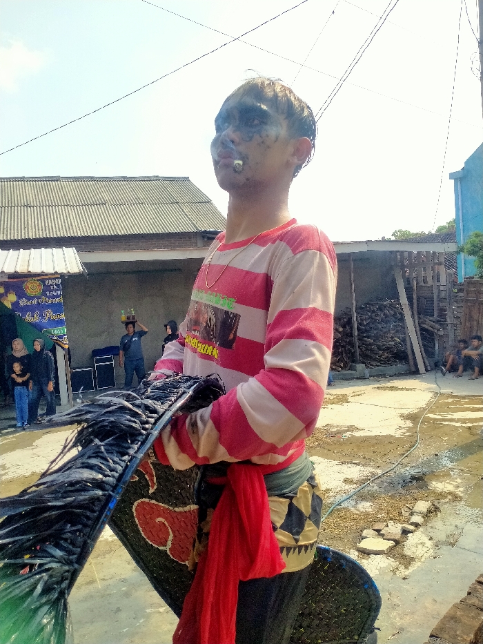 The image captures an outdoor daytime scene in Sepat, Indonesia. A young male, positioned centrally, faces right with his head slightly tilted upwards. He has dark, smudged makeup around his eyes and mouth, light-colored hair, and a lit cigarette in his mouth from which smoke emanates. He wears a long-sleeved shirt with wide alternating red/pink and white horizontal stripes, a gold-colored chain, a red sash tied around his waist over a dark and white patterned garment, and dark trousers. He holds a large, flat, dark object, possibly a prop, adorned with black fringe or tassels and a red, stylized cloud-like symbol.
The ground is unpaved and appears wet or muddy in sections, with a light-colored hose visible on the right. In the background to the left, a structure with a corrugated metal roof partially covers a banner displaying Indonesian text, including "Sepat" and "HARI HU...", confirming the city location. Several individuals, including women (one wearing a hijab) and a child, are visible observing near the banner. Two large black sound speakers are positioned near them. Further back, a man in a blue shirt carries a tray of items on his head. To the right, another building with a corrugated metal roof stands beside a large stack of cut wood. Two individuals are seated on the ground near the woodpile. A light blue building or wall is visible on the far right. The sky overhead is clear.