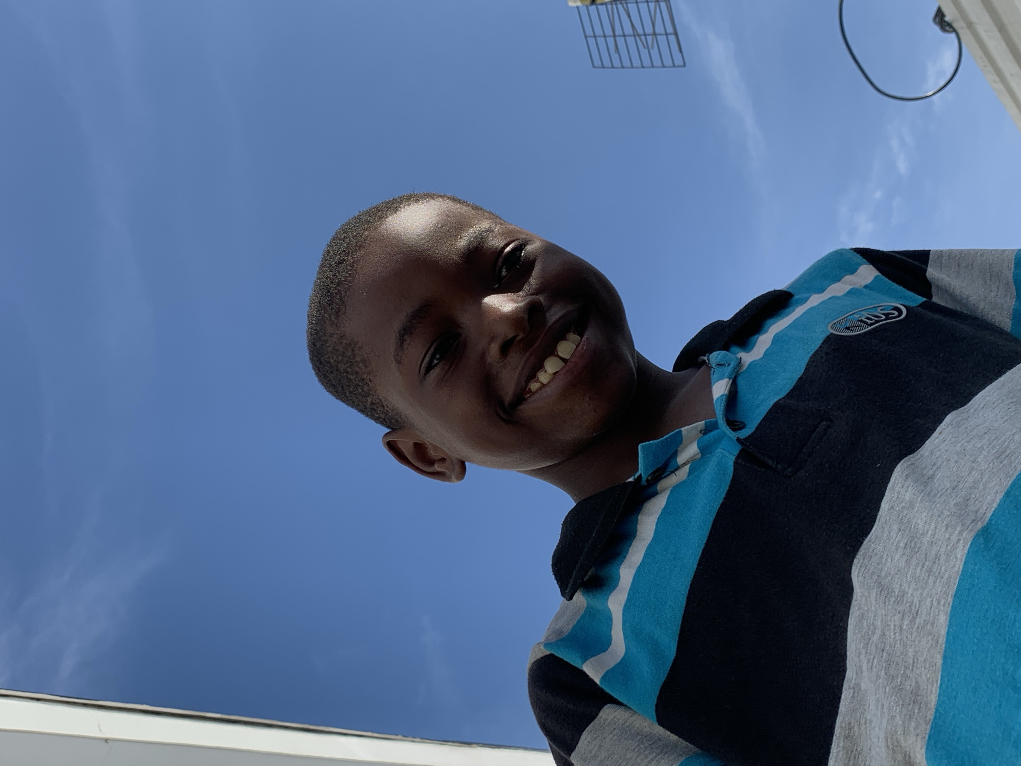 A low-angle outdoor portrait features a young Black boy looking up and smiling broadly at the camera. He is positioned in the lower-center of the frame, with his head tilted slightly. His face is illuminated, highlighting his wide, joyful smile that shows his teeth.
The boy is wearing a polo shirt with distinct vertical stripes in light blue, black, and grey. A small, oval-shaped emblem, possibly a logo or badge with indistinct text, is visible on the left chest area of his shirt.
The background is dominated by a clear, bright blue sky with subtle wispy white clouds. Due to the upward-facing angle, the sky fills most of the upper and mid-sections of the image. In the upper right corner, parts of a metal grid-like structure, possibly an antenna or part of a building fixture, and a metal ring are visible. A white, straight edge, likely a roofline or ledge, runs along the bottom left corner of the frame. The strong light and clear sky indicate it is daytime. The boy's expression conveys happiness and engagement. The scene is set in Ibasa, Nigeria.