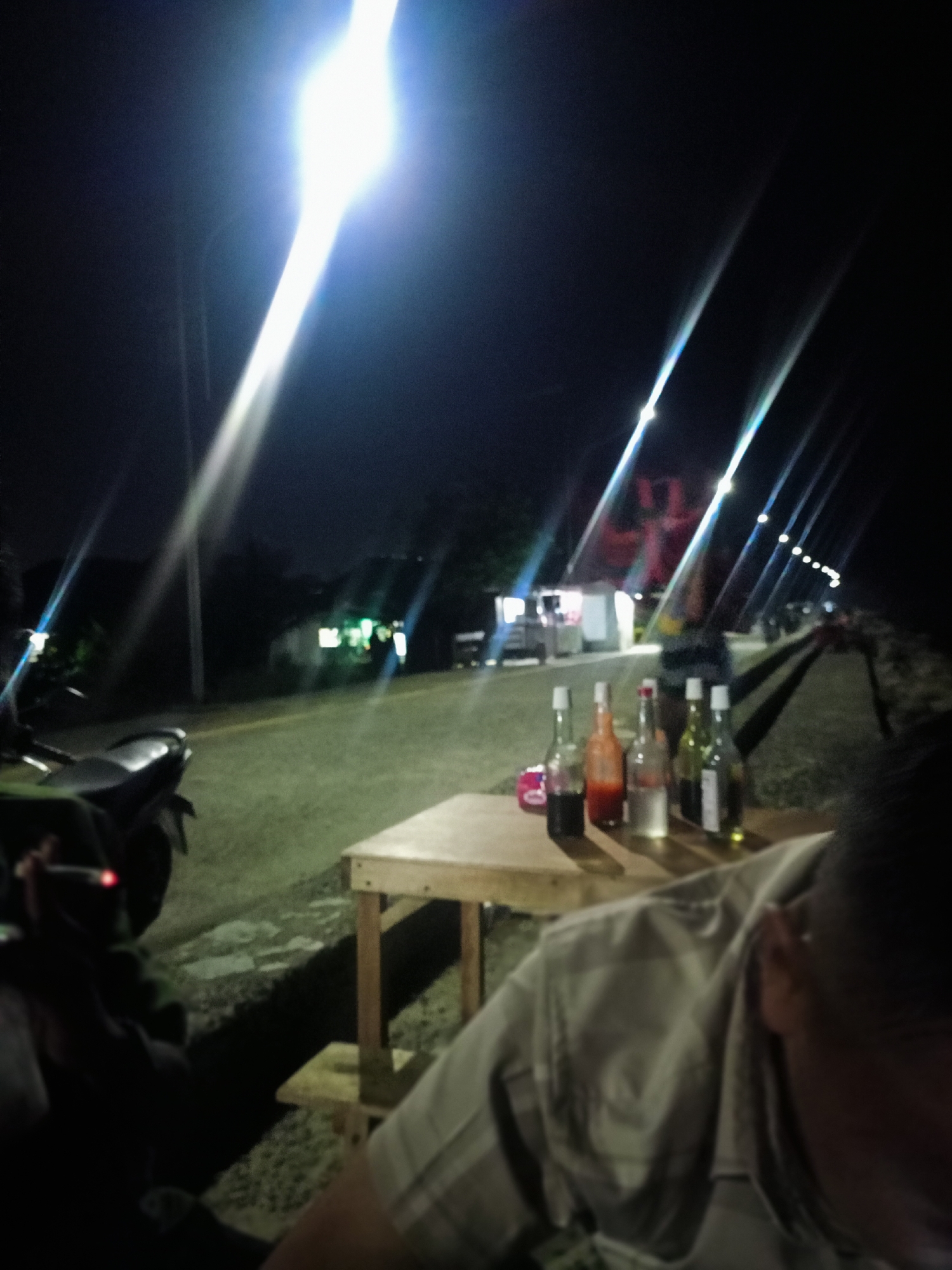 The image shows an outdoor scene at night in Lhokseumawe, Indonesia. A wooden table is centered in the foreground with a row of glass bottles on it; the bottles appear to contain different liquids. An individual sits in the foreground, partially visible, wearing a light-colored plaid shirt. To the left, someone is smoking near a parked motorcycle. In the background, there's a building illuminated by light. Street lights are visible along the roadside, causing bright light streaks in the image. There is a building with a red roof also illuminated by light.