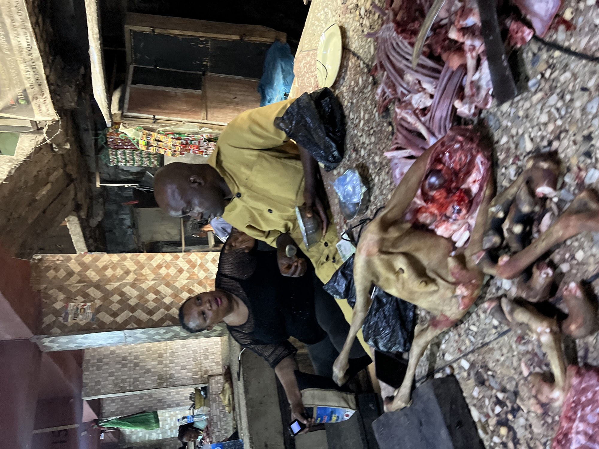 The image depicts a raw meat processing scene, likely in a market setting in Nkpor, Nigeria. Two individuals are visible: a man in a yellow shirt and a woman in a black shirt. They are standing near animal carcasses and various butchering tools. The carcasses include exposed ribs and internal organs. A black plastic bag and a metal container are near the meat. The environment appears to be a market stall, with tiled walls and other commercial goods visible in the background. An advertisement is visible with the text Head officer-A11/8 Upstal Nepior Main market, Branch Office:- A12/17 Kings & Queens Line Ogbo.