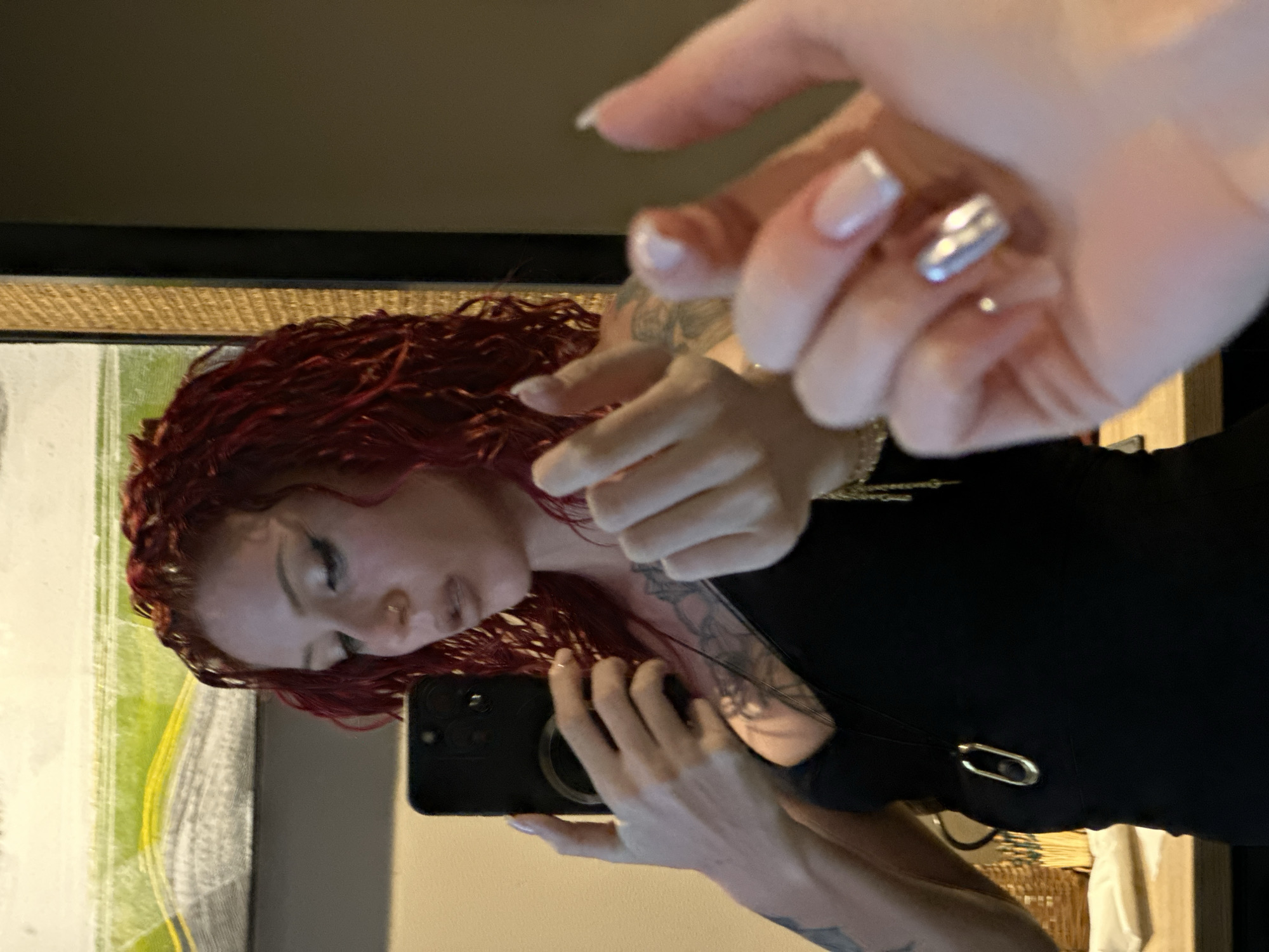A woman with red, curly hair and tattoos is taking a selfie in front of a mirror. She is wearing a black shirt and a necklace. Her nails are manicured, with one nail having a metallic silver design. The background includes a framed abstract painting and a wicker basket. The location is unknown, and there is no indication of it being in Indonesia.