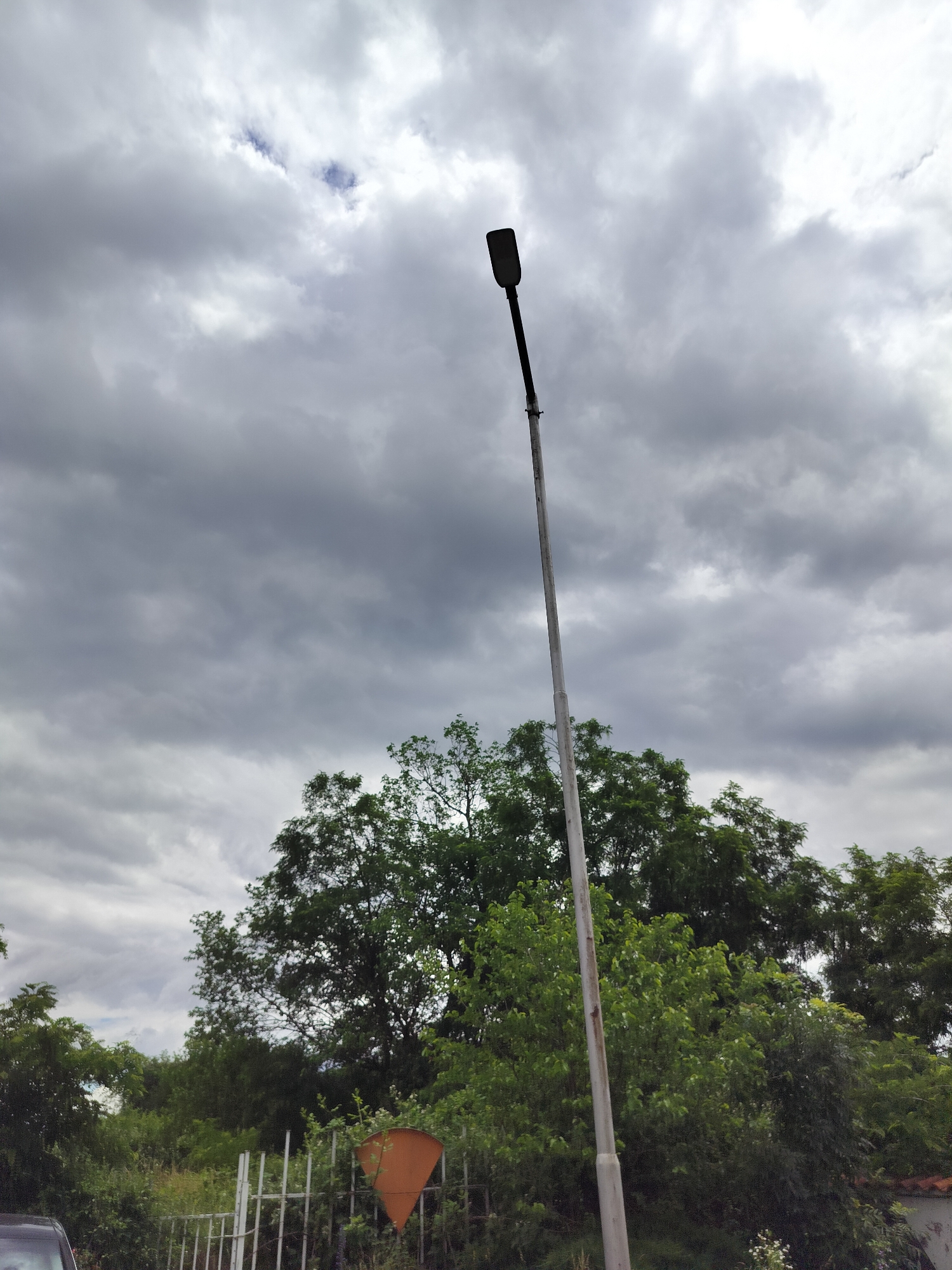 This is a ground-level, outdoor photograph of a street lamp against a cloudy sky. The street lamp, made of metal and painted a light color, stands tall and is positioned in the right-center of the frame. The lamp head is dark and rectangular. Below the lamp, a dense line of green trees forms the middle ground. In the foreground, to the left, a portion of a dark-colored vehicle is visible. To the right of the vehicle, a white metal fence runs horizontally, with a triangular orange sign attached to it. The background is dominated by a sky filled with heavy, gray clouds. The scene appears to be located in Tarnicheni, Bulgaria, based on the provided context.