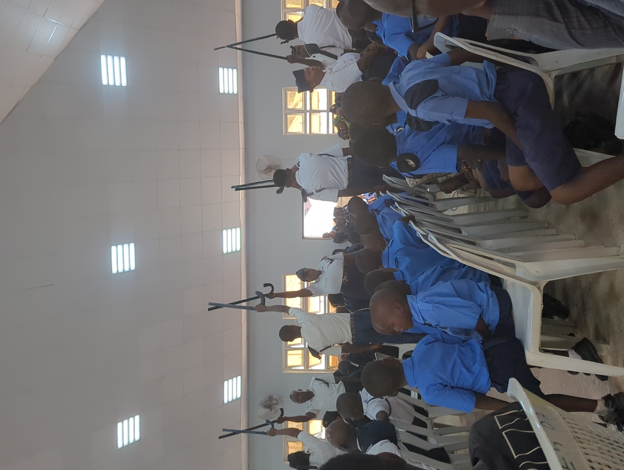 A large, brightly lit room with a white tiled ceiling containing recessed rectangular lights, holds several rows of seated individuals in blue uniforms with navy shorts, interspersed with white uniformed individuals, holding what appear to be black batons or sticks. Some seated individuals are in civilian clothes. The location is Sabon Tasha, Nigeria.