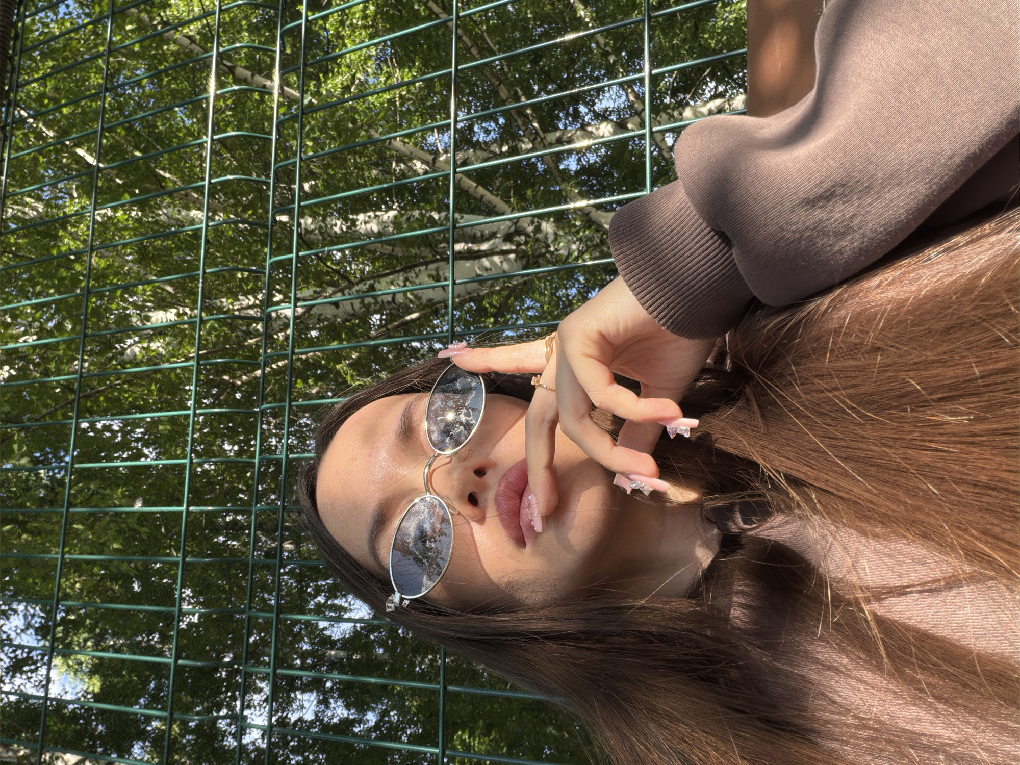 The image shows a young woman posing outdoors, captured from a slightly upward, close-up perspective. She has long, dark brown hair and is wearing a brown long-sleeved top or sweatshirt. Her left hand is raised near her face, featuring rings on her fingers and long, pink, manicured nails adorned with rhinestones. She is wearing round, dark-lensed sunglasses that reflect the surrounding trees and sky. Her lips are slightly parted and appear to have pink lip gloss.
Behind the woman, a green mesh fence creates a prominent grid pattern. Through the fence, a background of lush green trees is visible, including several birch trees with their characteristic white bark, suggesting a natural outdoor setting like a park or garden in Izhevsk, Russia. The bright sunlight indicates a clear, sunny day, likely late morning or afternoon, as evidenced by the strong shadows cast by the fence and on her face.
The original image is rotated ninety degrees counter-clockwise, giving the impression that the woman's head is tilted to the right. There is no visible text in the media file.