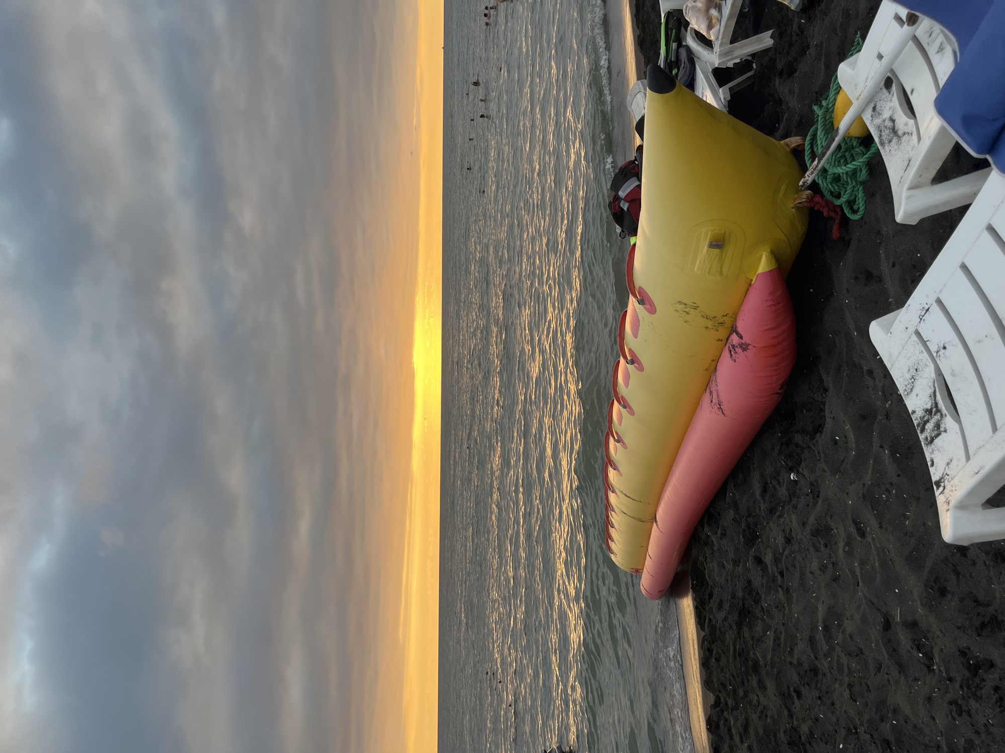 The image depicts a beach scene in Ureki, Georgia, at sunset. The sky is filled with dramatic clouds, ranging from dark grey to lighter hues, with a vibrant band of orange and yellow light illuminating the horizon where the sun is setting over the Black Sea. The sea itself is calm, with gentle waves lapping at the dark sand shore.
In the foreground, a large, inflatable banana boat lies partially on the black sand, positioned lengthwise. The banana boat is primarily yellow, with red accents, and a pink inflatable section attached to its underside. Nearby, on the sand, are several white plastic beach chairs, some of which appear to be occupied or have items on them. A green rope is coiled on the sand near the banana boat.
In the water, several people are visible swimming or wading. The overall atmosphere is one of a tranquil evening at the beach, with the warm colors of the sunset contrasting with the dark sand and the muted tones of the sky and sea.