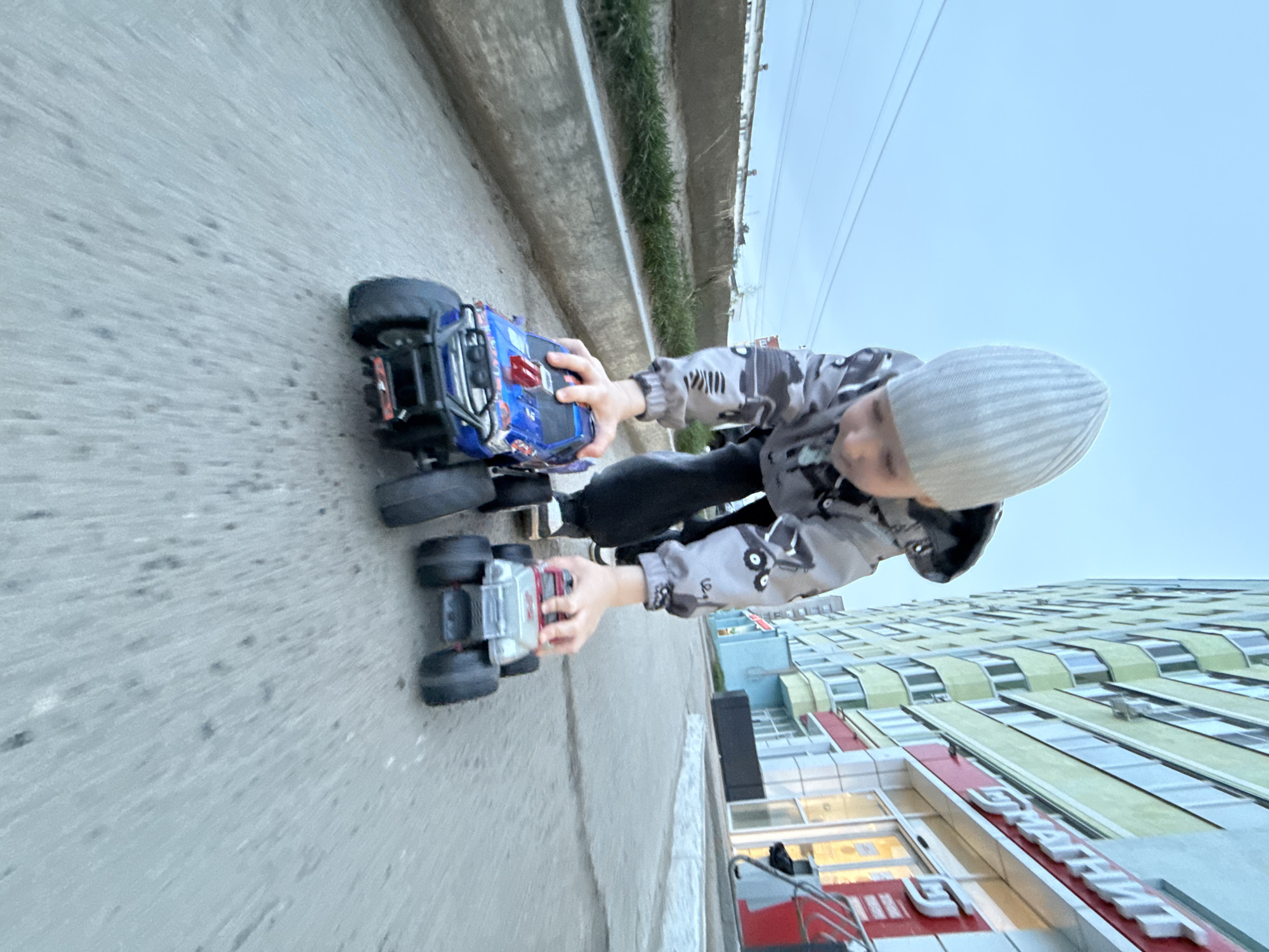 A child, wearing a gray patterned jacket and a light gray knit hat, is holding two toy monster trucks on a concrete sidewalk. The child is positioned between the sidewalk's edge, which is bordered by a concrete wall with grass growing along its edge, and a building. The building has multiple floors and features a sign with the Russian word "Магнит" visible, identifying it as a "Magnit" store. The sky is overcast. This scene appears to be taken in Chaikovsky, Russia.