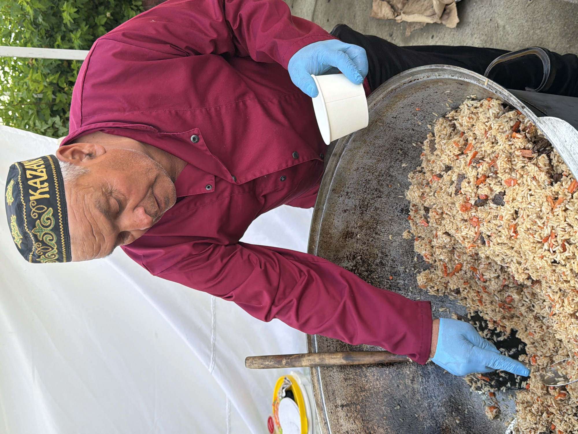 A man wearing a burgundy chef's coat, black pants, and a black and gold patterned skullcap with the word "KAZAN" embroidered on it is preparing a large pot of rice pilaf. He is wearing light blue disposable gloves. In his left hand, he holds a white paper cup, and his right hand, gloved, is reaching into the pilaf, which contains rice, pieces of meat, and carrots. A wooden spoon is also visible in the pot. The man is standing in front of a large, weathered metal pot. Behind him, a white tent or canopy is visible, along with some greenery. The ground appears to be concrete or asphalt. The scene suggests an outdoor food preparation setting.