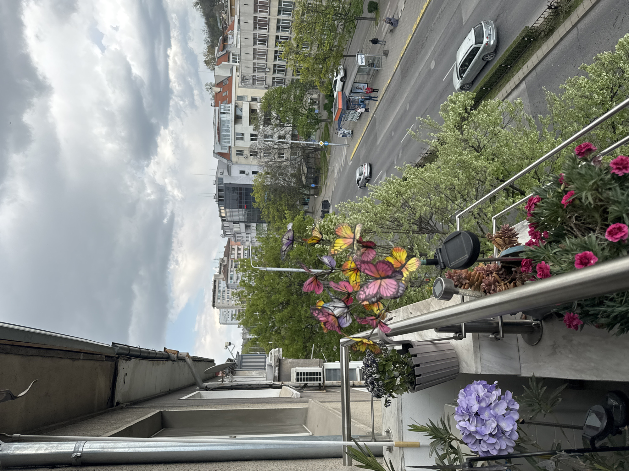 The image is an eye-level, upward, sideways shot taken from a balcony. A grey metal railing with plants and butterfly decorations lines the balcony. A potted purple hydrangea and other flowers are present. The view overlooks a street scene in Plovdiv, Bulgaria, featuring a road with cars, buildings, trees, a bus stop with people, and a street crossing. The sky above is cloudy.