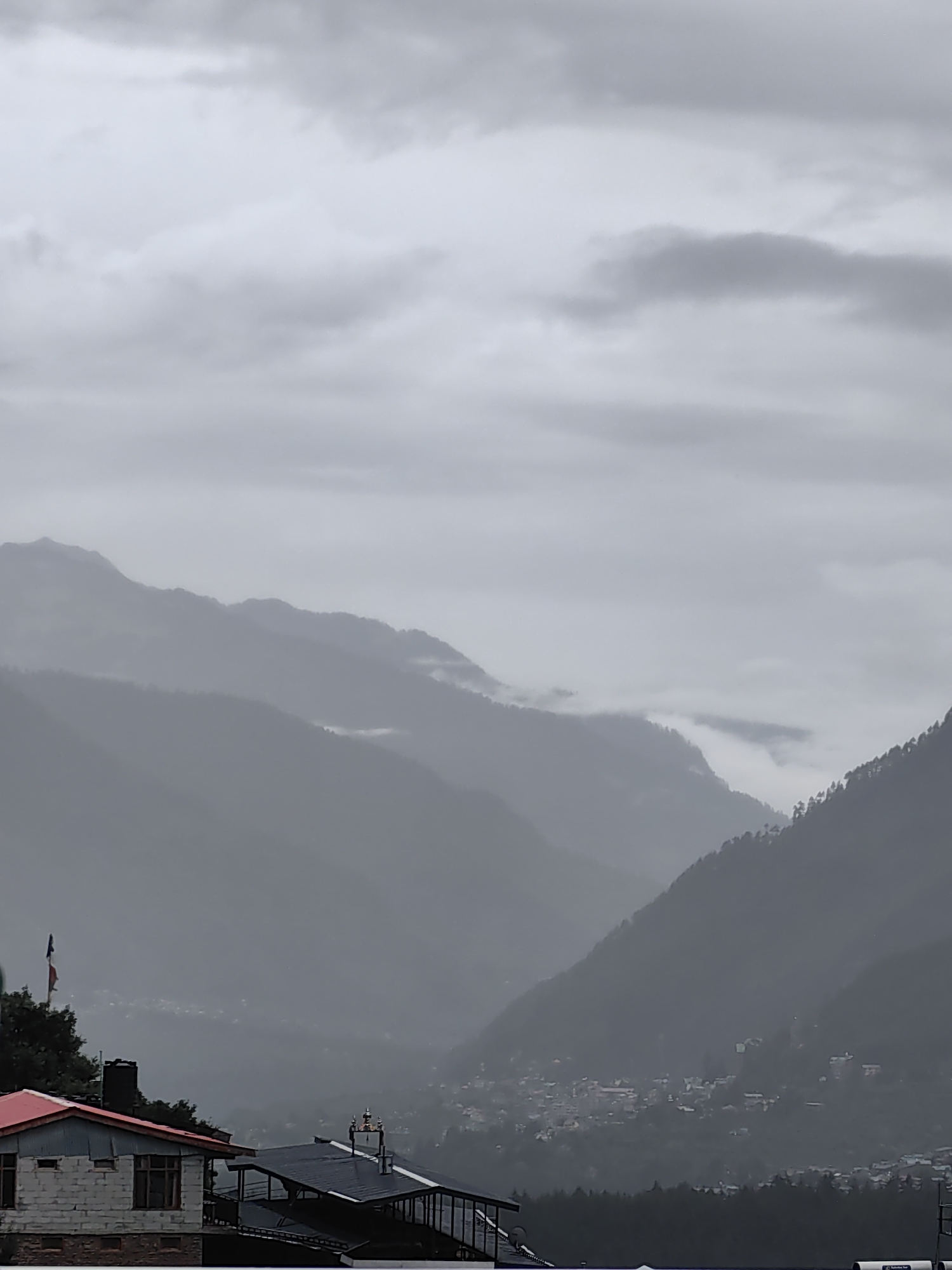 The image depicts a misty mountain landscape with a town nestled in the valley. In the foreground, there are several buildings with distinct roof structures. The building on the left has a red roof and white brick walls, with visible windows. To its right, another building features a dark, sloped roof. A flag on a pole stands prominently to the left of these buildings, adding a splash of color to the otherwise muted palette. The background is dominated by layered mountain ranges that fade into the mist and clouds, suggesting a high-altitude or overcast environment. A small settlement or town can be seen scattered across the valley floor, with houses appearing as small dots through the haze. The overall atmosphere is subdued, with soft, diffused light characteristic of a cloudy or rainy day in Vashisht, India, a location known for its hot springs and proximity to mountainous terrain.