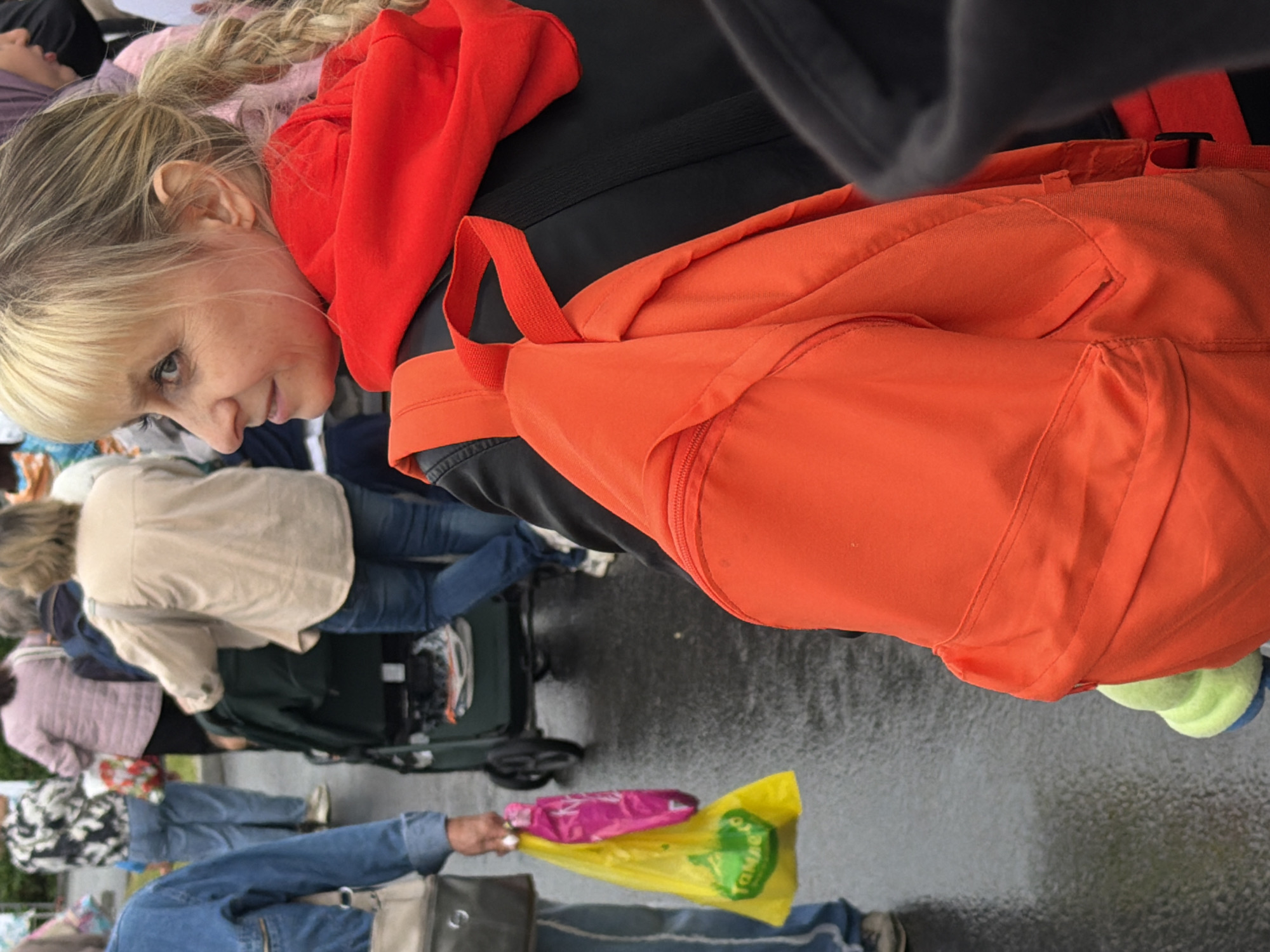 The image depicts a street scene with several people. A woman in the foreground is wearing a bright red hooded sweatshirt and a black jacket, with a large orange backpack visible. She has blonde hair pulled back in a braid. To her left, another person in a blue denim jacket is holding a yellow plastic bag containing a pink bag. In the background, other individuals are visible, some pushing a stroller or cart. The ground appears wet, suggesting recent rain. The location is identified as Unknown, Unknown.