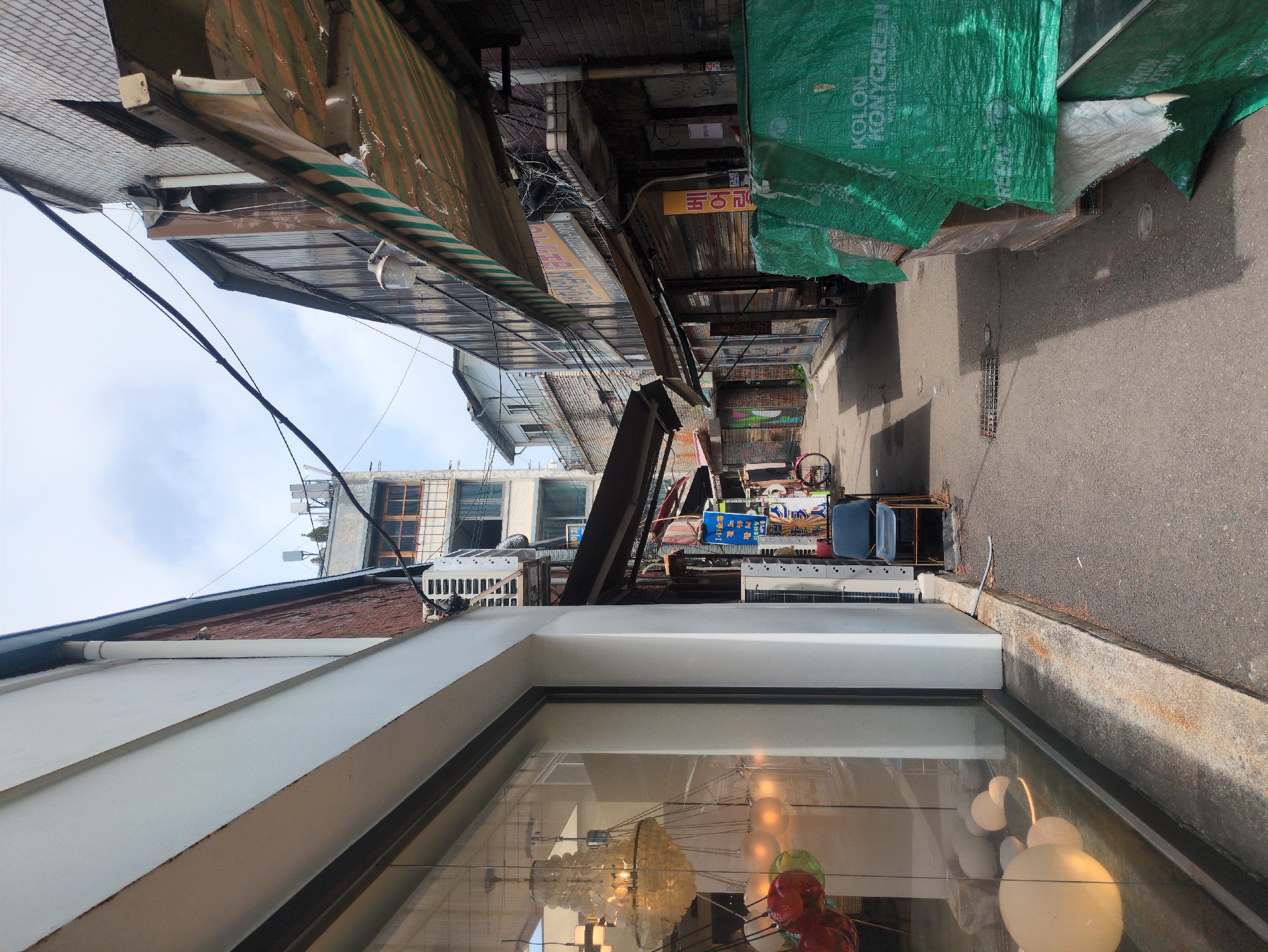 This image shows a narrow alley in Seoul, South Korea. The alley is lined with buildings of varying heights and architectural styles. There are numerous electrical wires running overhead. A green tarp covers some items on the right. The foreground features a building with large windows reflecting interior lighting fixtures. The alley appears cluttered with various objects, including what looks like merchandise for sale.