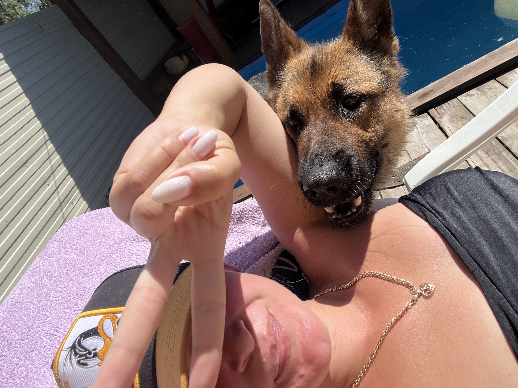 The image depicts a close-up of a woman and a German Shepherd dog. The woman is wearing a black top and a gold chain necklace with a heart pendant. She is also wearing a black baseball cap with a cartoon duck character on it and a purple towel is behind her. Her right hand is raised, obscuring part of her face, and she is making a peace sign. The German Shepherd is leaning into the frame, with its head resting on the woman's shoulder and its mouth open, revealing its teeth. The scene appears to be outdoors, possibly on a deck near a body of water, given the presence of wooden planks and a blue surface that could be a pool or lake. The background includes a grey corrugated metal structure. The lighting suggests it is daytime, with strong sunlight casting shadows. There is no specific information in the image that indicates a location in Novosibirsk, Russia.