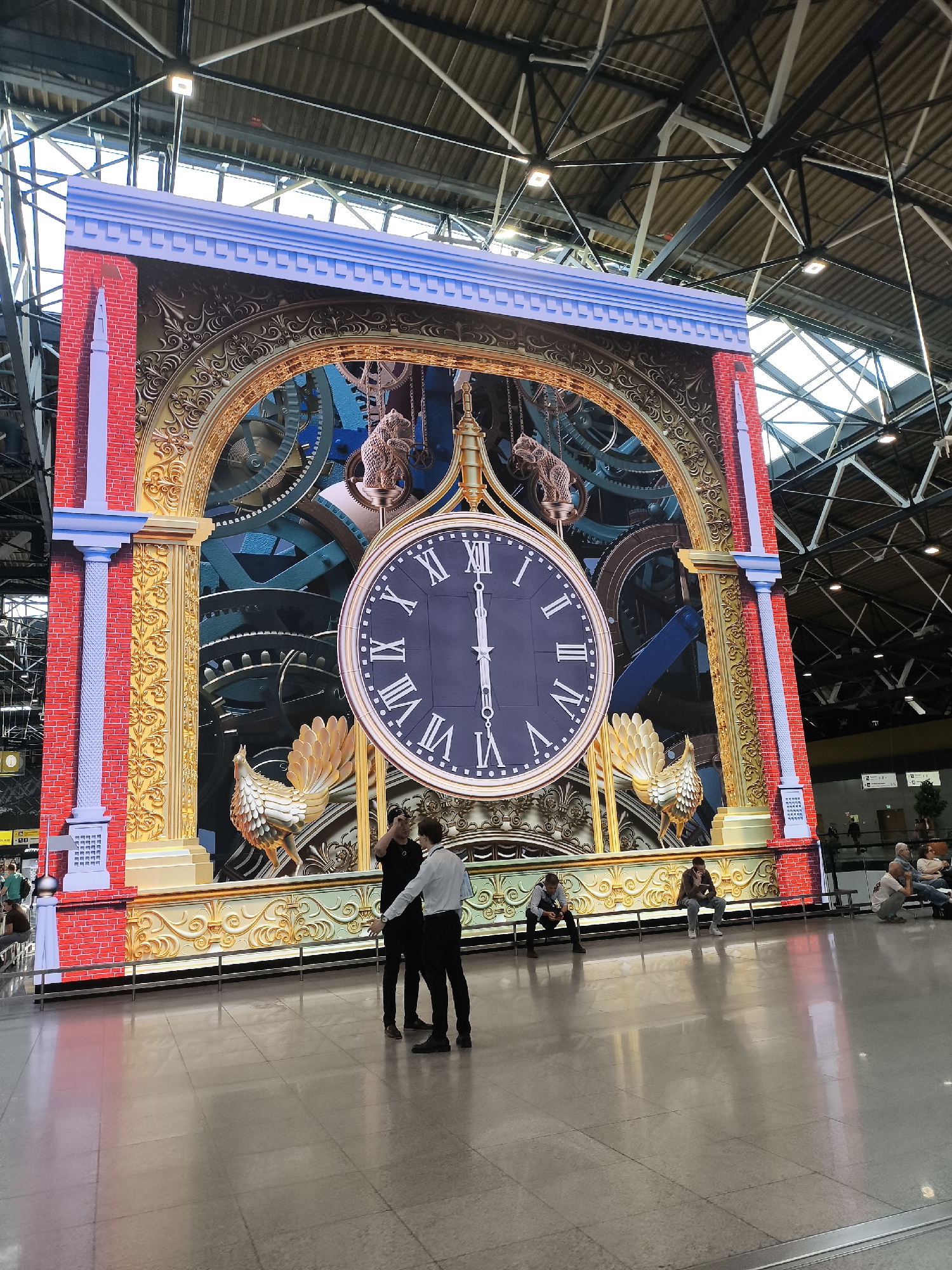 The image depicts a large, elaborate digital clock display prominently positioned within Sheremetyevo International Airport Terminal B in Khimki, Russia. The display itself mimics a monumental, ornate mechanical clock, featuring a central dark blue clock face with white Roman numerals indicating XII o'clock (12:00), with both hands pointing upwards. Surrounding the clock face is a golden, intricately carved archway and framework, suggesting a steampunk or classical aesthetic. Behind the clock face and within the arch are visible large, dark blue and bronze gears and mechanisms. The display is framed by faux red brick columns with light blue architectural accents at the top and base, including pilasters and cornices. A golden rooster figurine stands on a lower ledge on the left side of the clock, mirrored by a similar, less visible figure on the right. A small, animal-like figurine (possibly a bear or monkey) hangs from chains above the clock on the left.
The setting is a spacious, brightly lit airport terminal. The floor is composed of reflective light-colored tiles, showing clear reflections of the display and individuals. The ceiling is very high, revealing exposed metal trusses, girders, and skylights providing natural light, supplemented by square artificial light fixtures mounted on the structure.
In the foreground, two men stand facing each other, seemingly engaged in conversation. One man, wearing a black shirt and black pants, gestures with his left hand. The other man wears a white long-sleeved shirt and dark pants, holding a light-colored item in his left hand. Further in the mid-ground and background, several other individuals are visible. Some are seated on metallic benches, looking at mobile devices or resting, while others are standing or walking. Their attire is casual, consistent with travelers. The overall atmosphere suggests a quiet period in the terminal, with moderate pedestrian traffic. Visible signs and further architectural elements of the terminal are present in the distant background.
