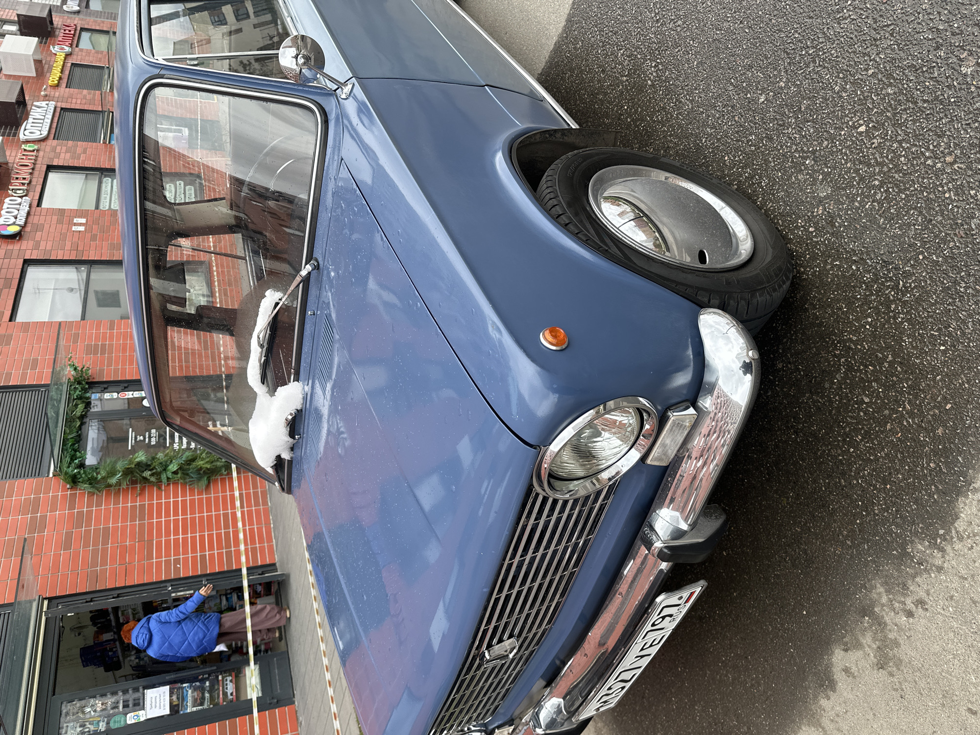 The image presents a street scene, likely in Moscow, Russia, featuring a light blue vintage car parked beside a building with red brick facades. The car, which appears to be a VAZ-2101 (Lada 1200) or a similar model, has chrome bumpers, grille, and headlight bezels. A small amount of snow is visible on its windshield, indicating cold weather, and the asphalt street is wet. A notable feature of the car is a spare tire with a chrome hubcap mounted vertically on its front right side.
A person dressed in a blue quilted jacket and brown pants is seen at the entrance of one of the storefronts, possibly entering or exiting. The building houses various businesses, identified by Cyrillic signs. These include "СОЦИАЛЬНАЯ АПТЕКА" (Social Pharmacy), "ОПТИКА" (Optics), and "ФОТО РЕМОНТ" (Photo Repair/Services), which also offers "ФОТО НА ДОКУМЕНТЫ" (Photos for Documents). Another sign displays "Требуется" (Wanted/Required) along with operating hours "10:00-22:00". The car's Russian license plate, "B627YE797 RUS", is visible, with "797" being a Moscow region code. The scene takes place during daylight hours, under what appears to be an overcast sky.