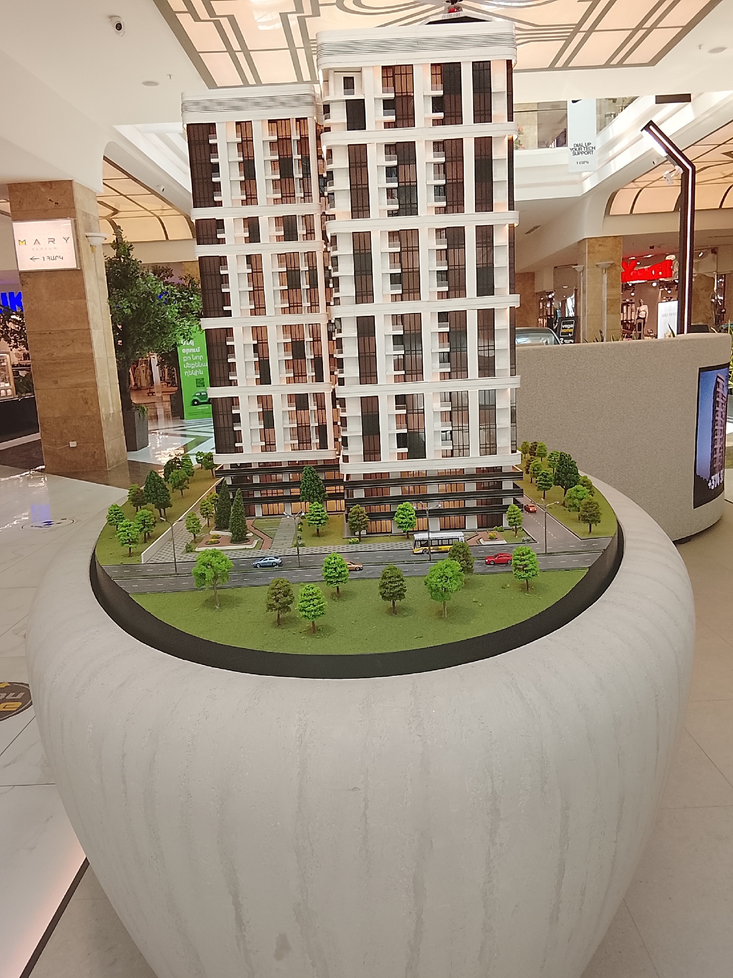 A large architectural model of two modern high-rise residential buildings is displayed prominently in what appears to be a shopping mall or large commercial space. The model features two tall, multi-story structures with white facades and numerous dark, recessed windows and balconies. Surrounding the buildings is a detailed miniature landscape including green grass, various types of trees, streetlights, and a road. On the road, there are several miniature vehicles: a blue car, a red car, and a yellow bus. The entire model is situated on a circular, dark green base, which is then set into a large, curved, textured white planter-like structure that sits on a tiled floor.
The background reveals typical mall elements: high ceilings with intricate geometric patterns and recessed lighting, large pillars covered in what appears to be marble, and other commercial displays. On the left, a sign reads "MARY" with "TERRAUN" below it, and an arrow pointing left with the Armenian text "1 ՅԱՐԿ" (likely "1st Floor"). Further back, a green banner with Armenian text, "օրում քո նոր մեքենա ոճին," is visible. In the background to the right, a sign with "DIAL UP YOUR TECH SUPPORT" is partially seen, and a sleek, modern light fixture stands near a dark store entrance. An LED screen embedded into the right side of the model's white base shows part of a phone number, "+374 9...". The scene is well-lit, suggesting daytime or bright indoor illumination. There are no people visible in the immediate vicinity of the model. The overall impression is a detailed property development advertisement within a public commercial environment.