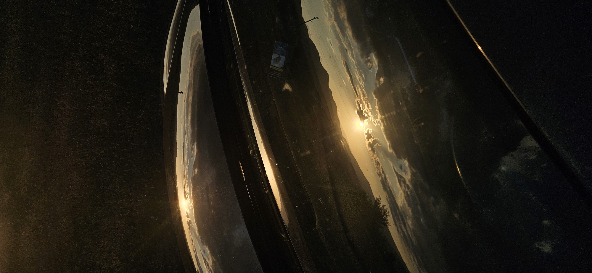 A close-up, distorted view of a sunset reflected on a dark, possibly metallic surface. The sun is positioned near a mountain, with some clouds. The image might be taken inside a vehicle. The location is not discernible from the image alone. There are no people visible.