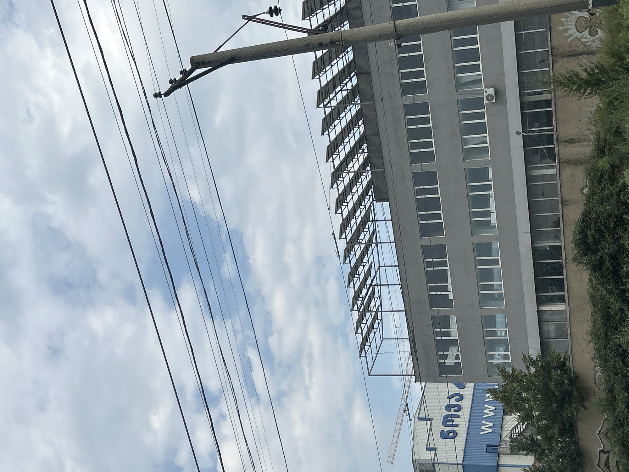 The image depicts an urban industrial scene with a multi-story gray building prominently featured, its facade punctuated by numerous windows. Above the building, a row of solar panels is installed on a metal frame. In the background, a blue building with white lettering, "WWW," and another blue structure with Georgian script, "6m33," are visible. A construction crane extends from behind the blue buildings. Power lines crisscross the sky, supported by a concrete utility pole. The foreground consists of dense green foliage and a low wall with graffiti. The scene is set against a cloudy sky.