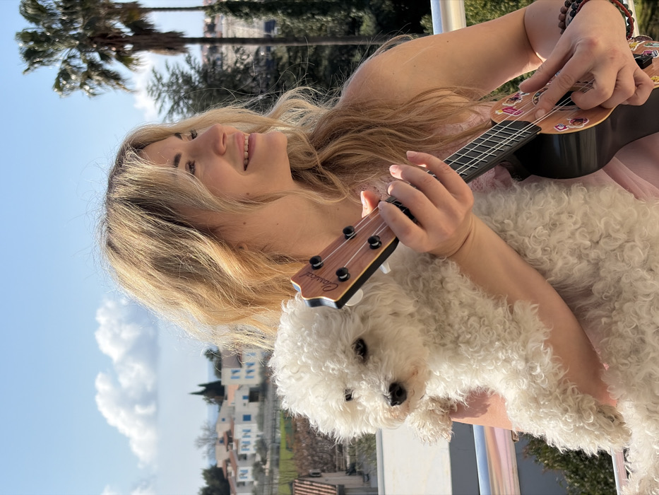A blonde-haired female is positioned outdoors on a balcony, holding a small, white, curly-furred dog in her left arm while her right hand grips and is positioned to play a ukulele. The ukulele features a "Classical" branded headstock and a light-colored soundboard adorned with multiple colorful stickers. The female, wearing a light-toned, possibly pink, sleeveless top and a multi-strand beaded bracelet on her right wrist, smiles and looks towards the viewer's right. The dog, a Bichon Frise type, faces the camera with a neutral expression. A silver balcony railing is visible in the foreground. In the background, under a clear blue sky with scattered white clouds, are white-painted buildings with terracotta roofs, characteristic of Mediterranean architecture, interspersed with green trees and vegetation. Two prominent palm trees are visible on the right side of the background. This architectural and natural environment is consistent with the city of Budva, Montenegro.