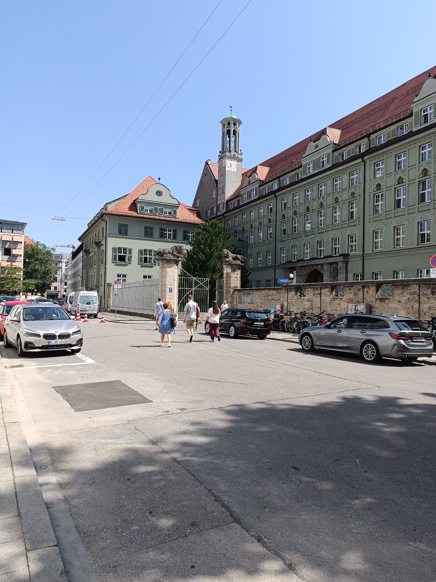 The image depicts a street scene in Munich, Germany, with a large, light green, multi-story building dominating the right side of the frame. The building features numerous white-framed windows and a reddish-brown tiled roof. To the left of this building, a smaller, light green building with a similar tiled roof is visible, featuring green shutters on some windows. A tall clock tower with a green, domed roof and a weather vane is attached to the larger building, near its left edge.
In the foreground, a street runs diagonally across the image. Several cars are parked or driving on the street. A silver BMW station wagon is prominent in the lower right quadrant, facing away from the viewer. A black BMW sedan is parked on the left side of the street. Other vehicles, including a white van and a red car, are visible further down the street.
A group of four pedestrians is walking across the street from left to right. They are casually dressed. A stone wall with a metal fence runs along the front of the larger building, separating it from the street. Two stone lion sculptures are positioned on top of pillars flanking an entrance within the fence. A blue sign with the word "POLIZEI" (Police) is mounted on the wall.
The environment appears to be an urban setting on a sunny day, indicated by the clear blue sky and the shadows cast by trees. The street is paved, and a sidewalk is visible on the left. Orange traffic cones are placed on the street near the white van. The overall impression is that of a typical city street with historical architecture. The specific location is near the entrance to the former Munich Police Headquarters at Ettstraße 2, 80331 Munich, Germany, which is now part of the State Attorney's Office.