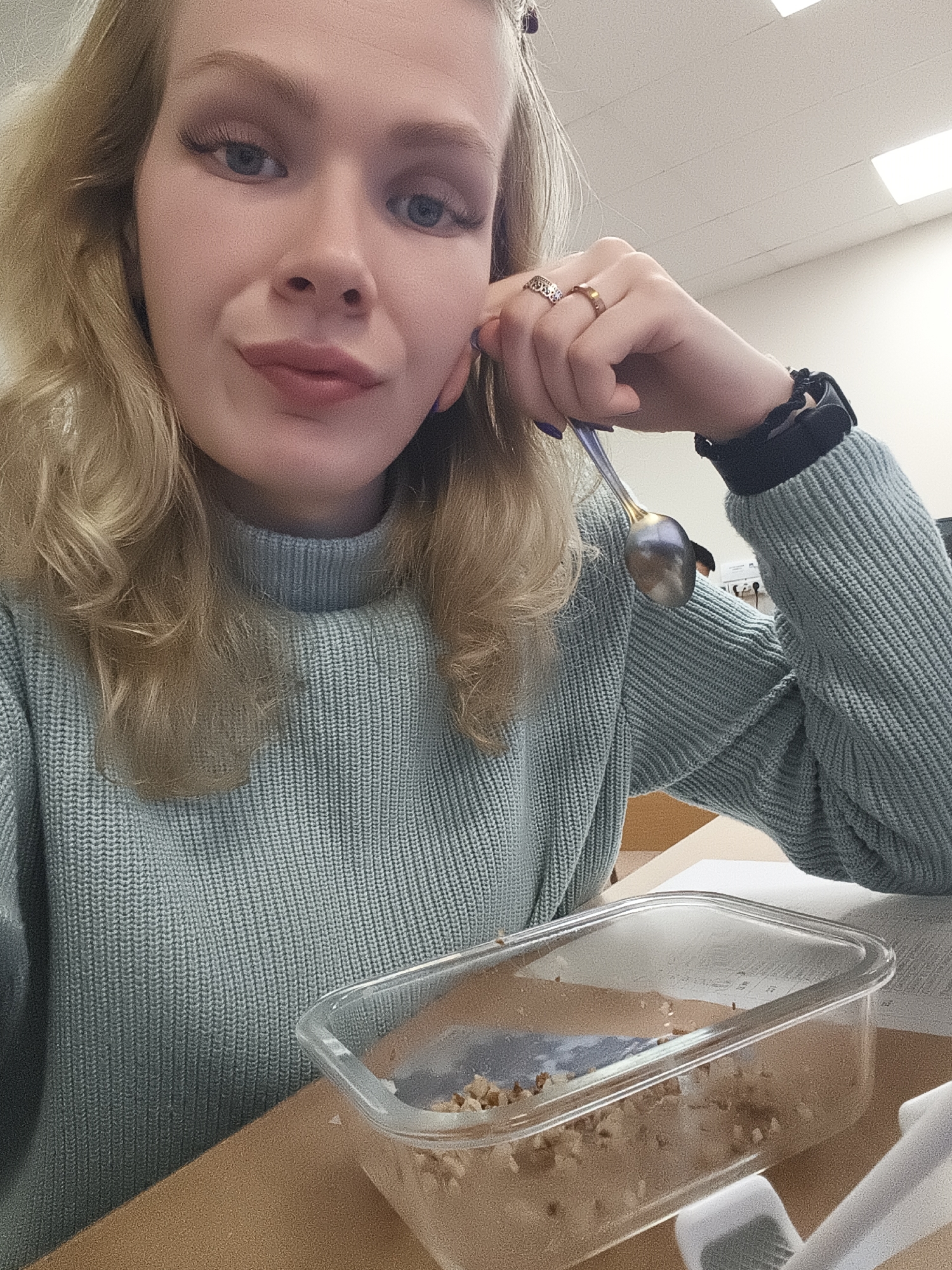 A woman is shown in a close-up selfie, seated at a desk. She has light blonde hair, blue eyes, and is wearing a light blue turtleneck sweater. A metal spoon is visible in her right hand, which is resting near her face, adorned with two rings. On her left wrist, she wears a black scrunchie or hair tie. A clear plastic container sits on the desk in front of her, containing what appears to be food with some brown liquid and solid particles. The background features a light-colored wall, a ceiling with fluorescent lights, and what seems to be an office or institutional setting. The city and region are unknown.