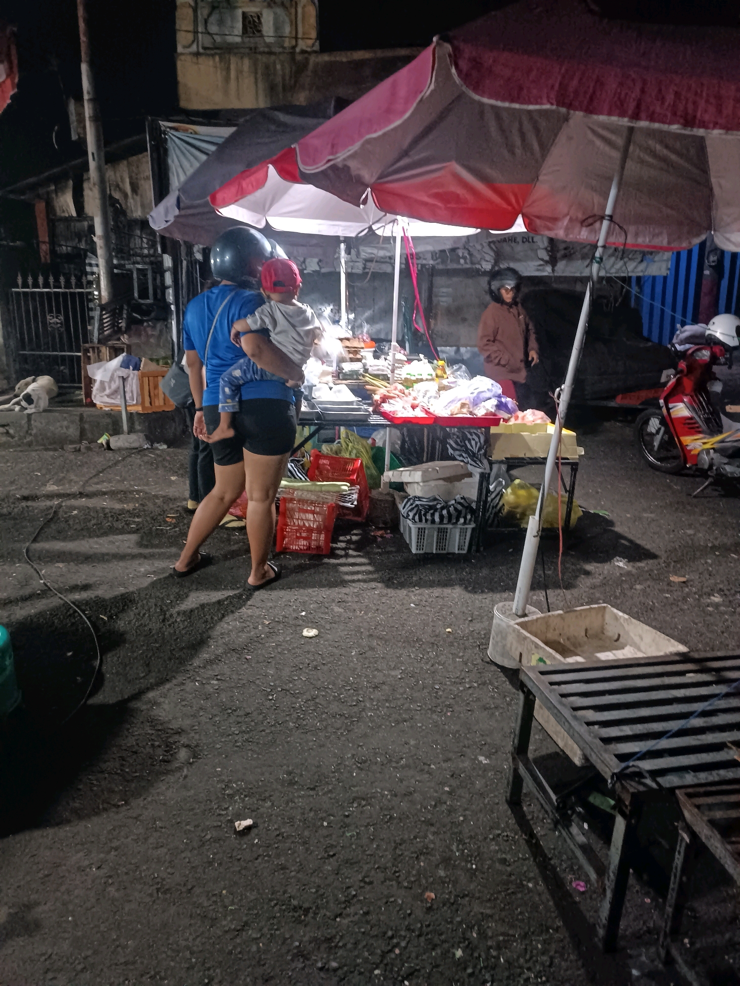 A nighttime street scene in Tabanan, Indonesia, captures a vendor stall under a large, multi-colored umbrella, displaying various food items on tables. In the foreground, a person in a blue shirt and black shorts carries a child. Another person, wearing a brown jacket, stands near the stall. A red scooter is parked to the right. The ground is paved and appears wet, with shadows cast by the stall and an umbrella. The background shows buildings and a metal fence.