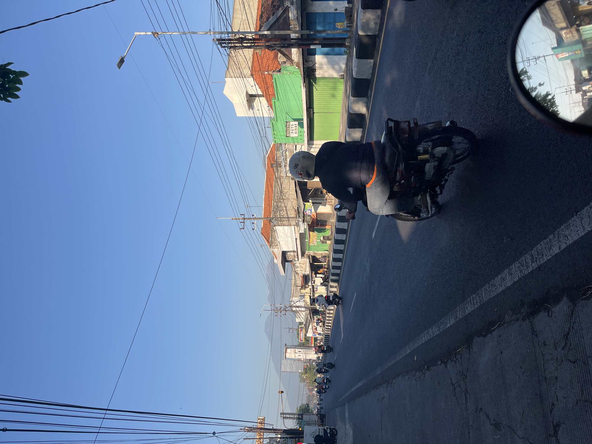A bright, clear day is depicted on a wide, asphalt road in Mindi, Indonesia, likely in the morning given the soft sunlight. The road is bordered by a sidewalk with a low concrete barrier. On the right side of the road, there are several buildings with commercial storefronts, some with red tile roofs and others painted green or white. A street lamp is visible on the right. On the left side of the road, more buildings and power lines are present. In the distance, mountains are visible through the haze.
The foreground features a motorcycle rider from behind, wearing a helmet and a dark jacket over a grey shirt and jeans with an orange waistband. The motorcycle is black and appears to be an older model. The rider is positioned in the center lane of the road, moving forward. Several other motorcycles are traveling in the same direction, further down the road, with riders also visible. In the reflection of the motorcycle's rearview mirror, a portion of the buildings and power lines across the street can be seen, along with a sliver of the bright blue sky.