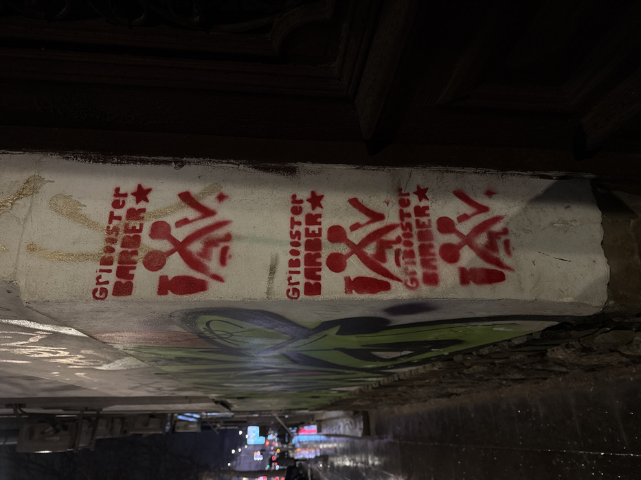 Three red "Gribooster Barber" stencils are painted on a white surface. Each stencil includes crossed scissors, a razor, and a star. The white surface appears to be part of a concrete structure, possibly beneath an overpass, with additional graffiti below. The background suggests an urban environment, with streetlights and city lights visible in the distance. The image is taken from a low angle, emphasizing the underside of the structure.